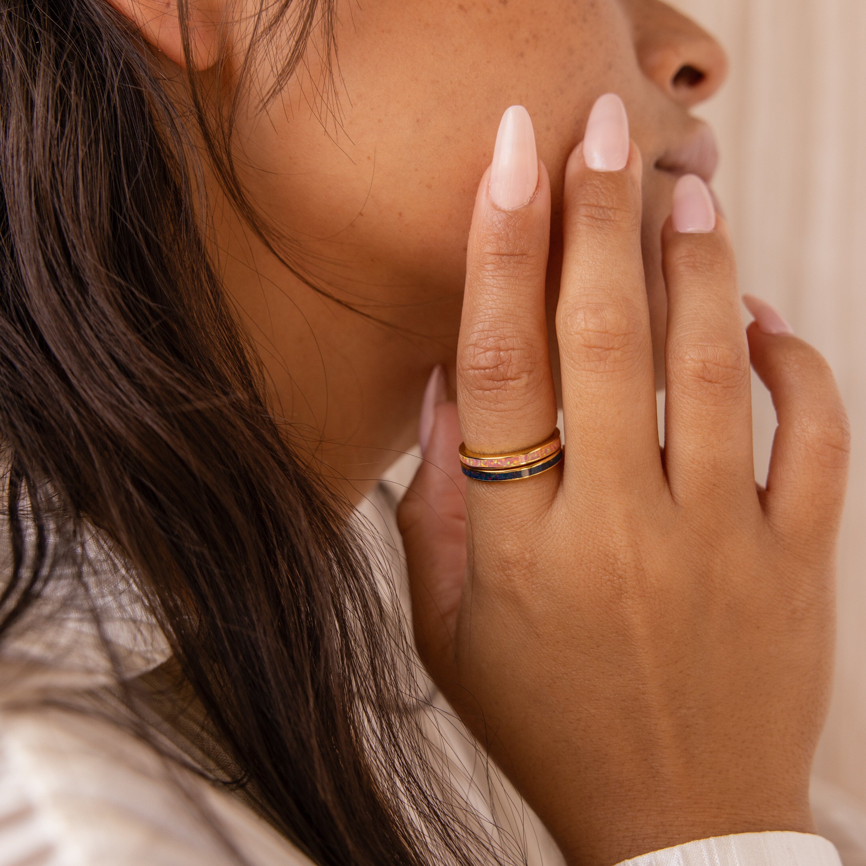 A woman with long nails touches her face, showcasing stacked rings on her finger, including the striking Indigo Opal Inlay Band.