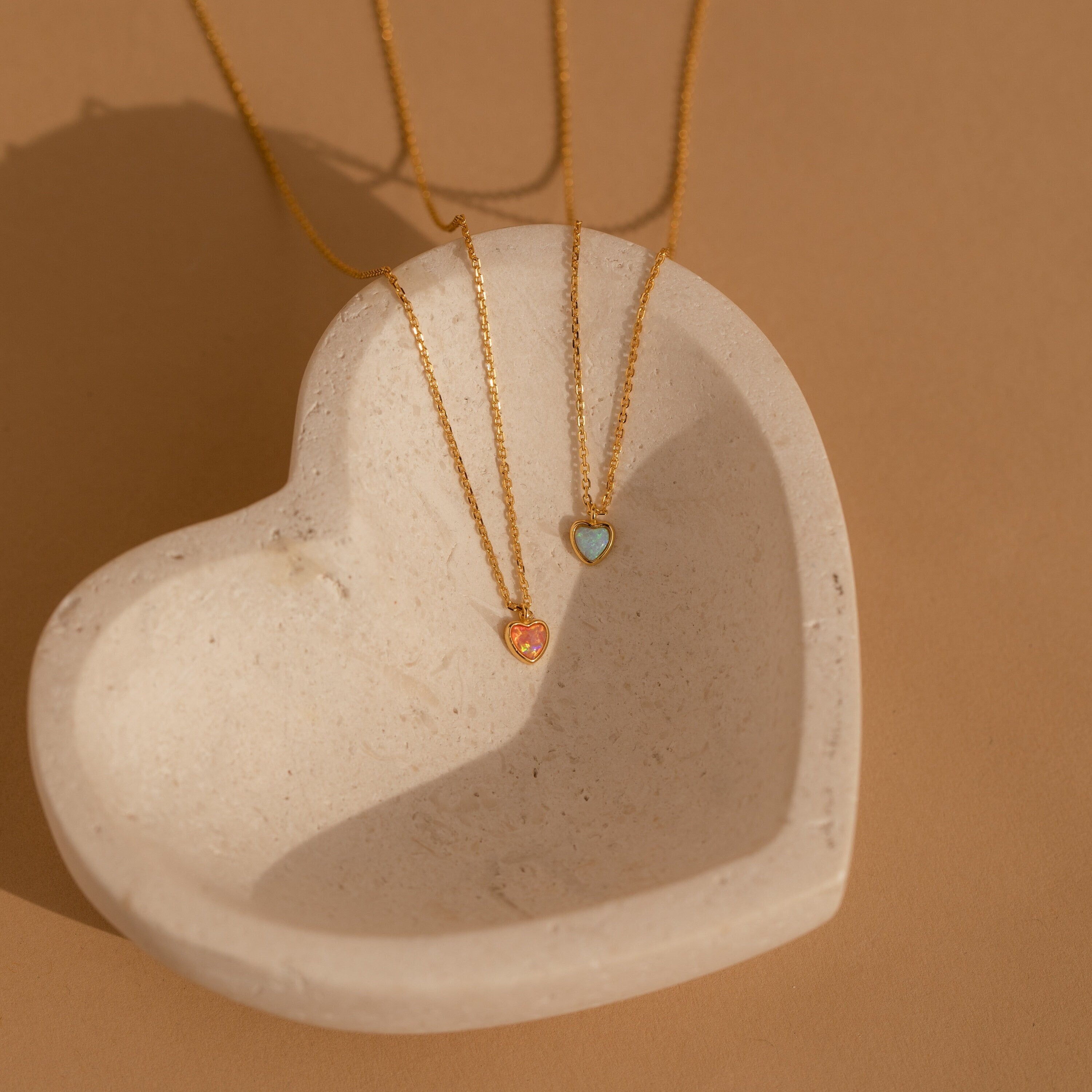 A Pink Opal Heart Necklace and a blue heart-shaped necklace rest in a cream-colored heart dish—an elegant way to display opal jewelry.