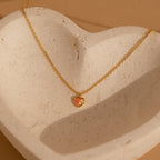 Gold necklace with a fine chain featuring a small heart shaped pink opal pendant, displayed on a textured stone dish.
