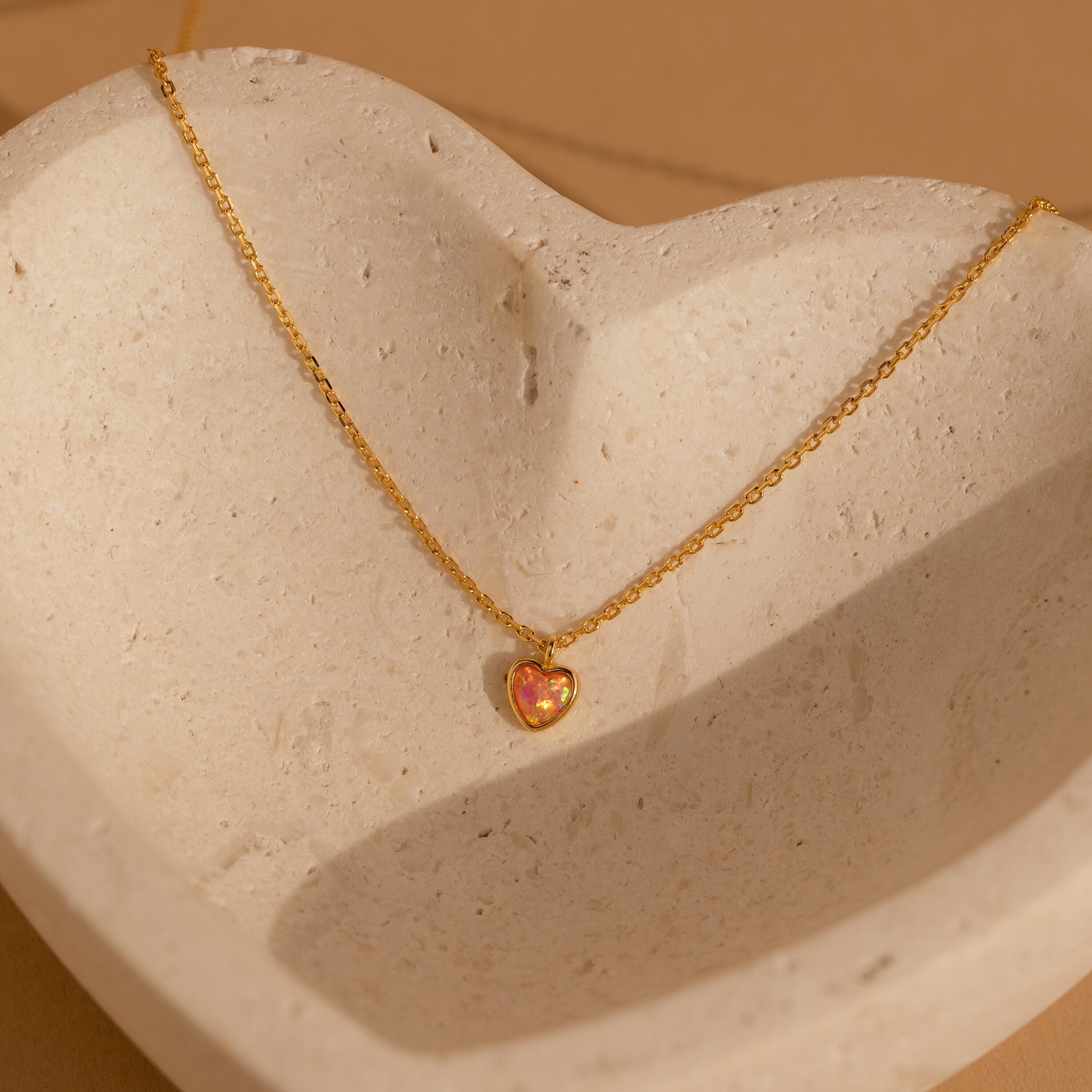 Gold necklace with a fine chain featuring a small heart shaped pink opal pendant, displayed on a textured stone dish.
