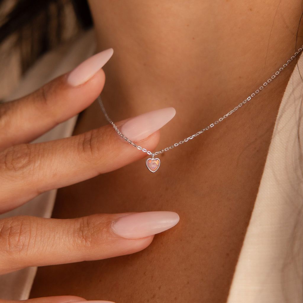 A hand with long pale-pink nails touches a delicate Pink Opal Heart Necklace, which rests gracefully on a woman's neck.