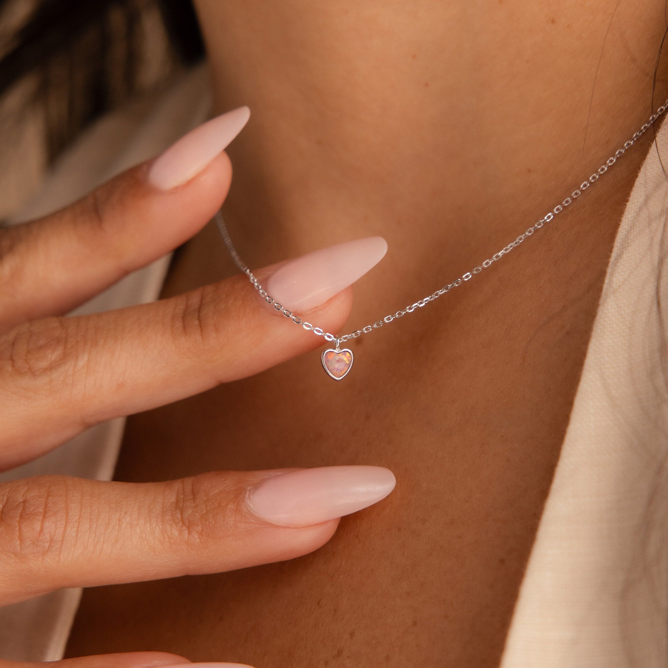 A hand with long pale-pink nails touches a delicate Pink Opal Heart Necklace, which rests gracefully on a woman's neck.