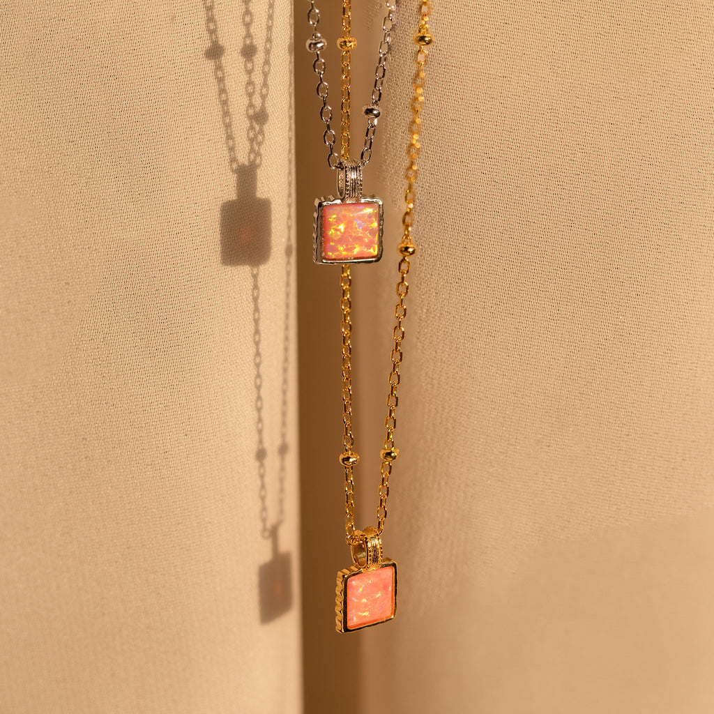 Two minimalist Pink Opal Pendant Necklaces, one in silver and one in gold, hang on satin satellite chains against a soft beige fabric background.