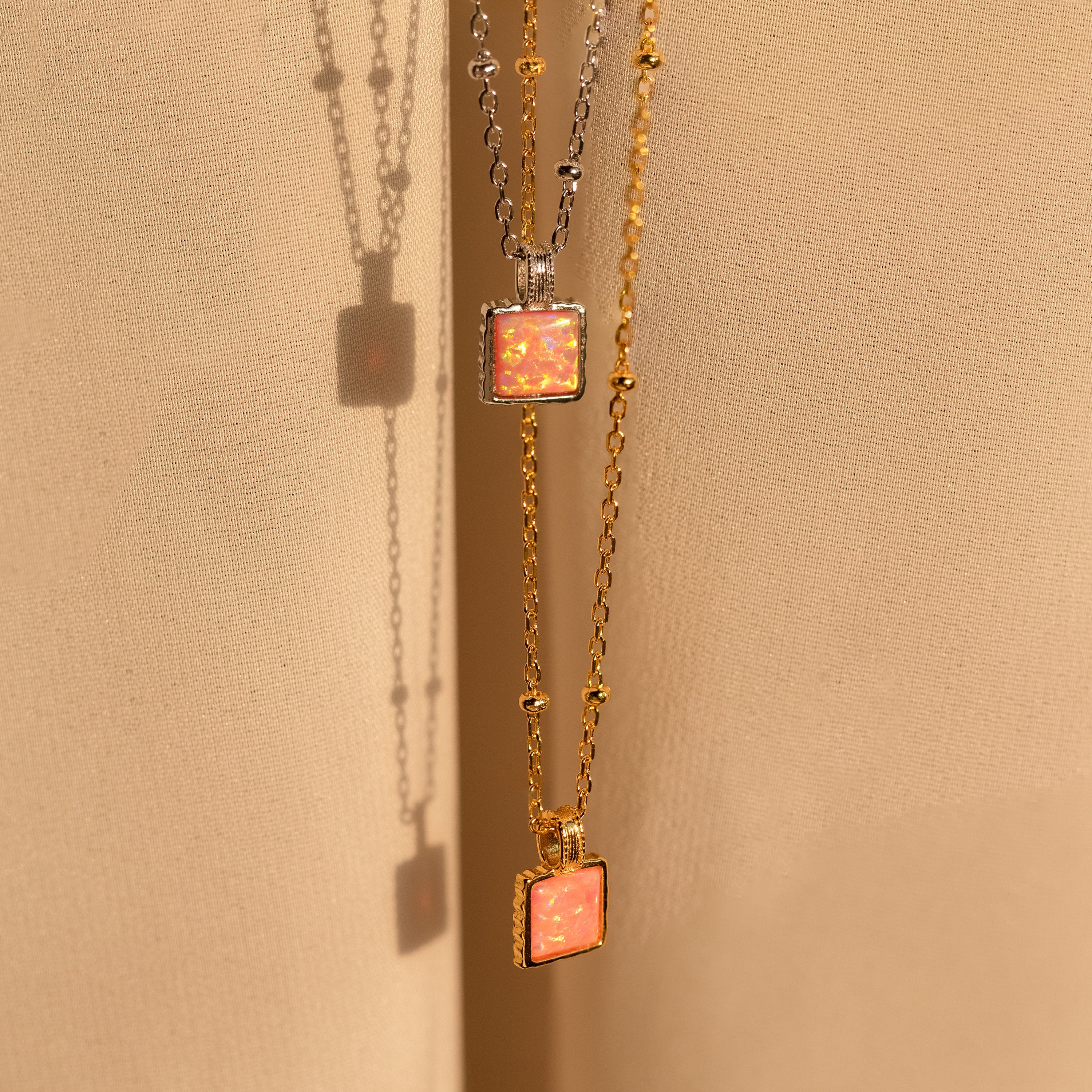 Two minimalist Pink Opal Pendant Necklaces, one in silver and one in gold, hang on satin satellite chains against a soft beige fabric background.