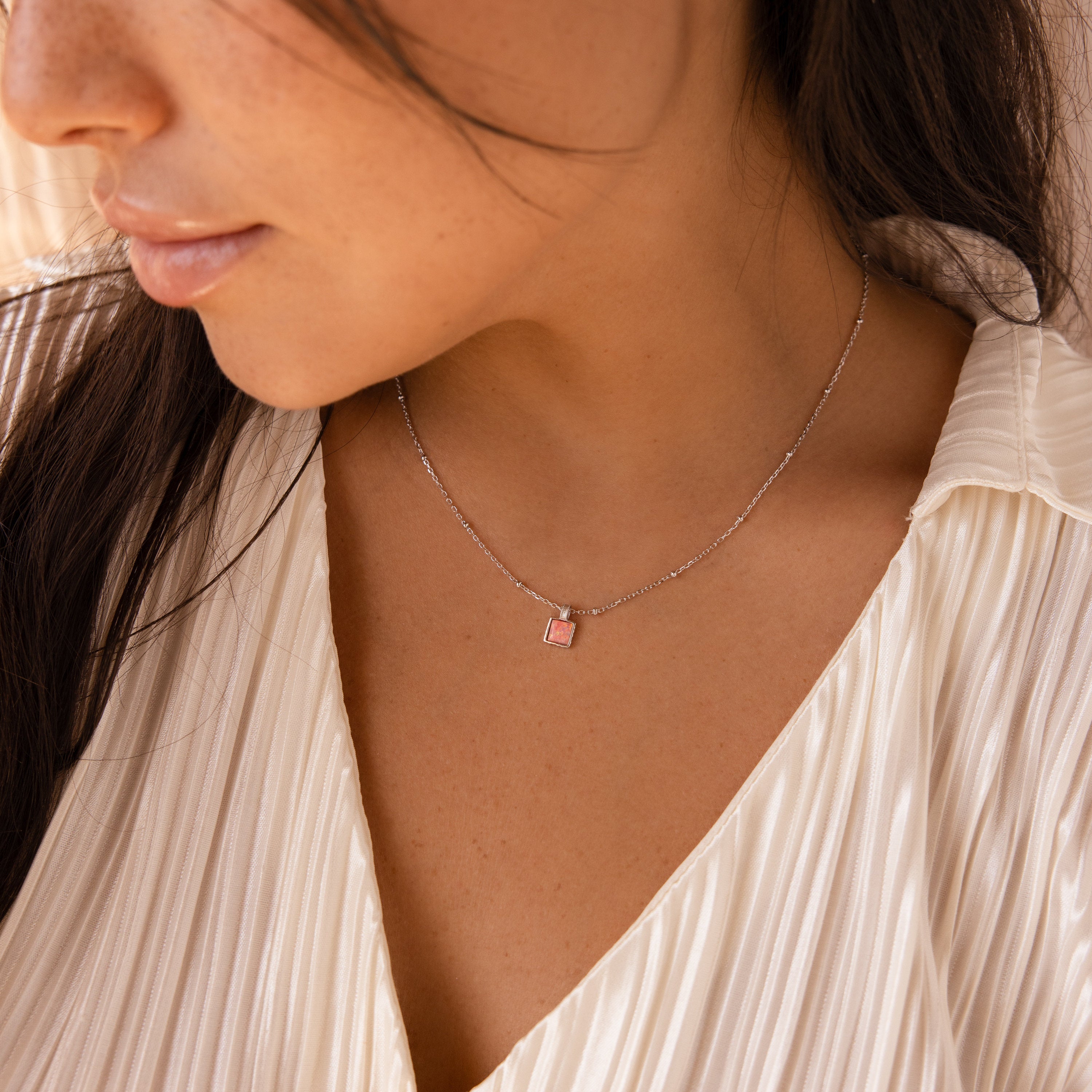 A woman in a pleated cream blouse wears the Pink Opal Pendant Necklace—a delicate satellite chain with a pink rectangular pendant.