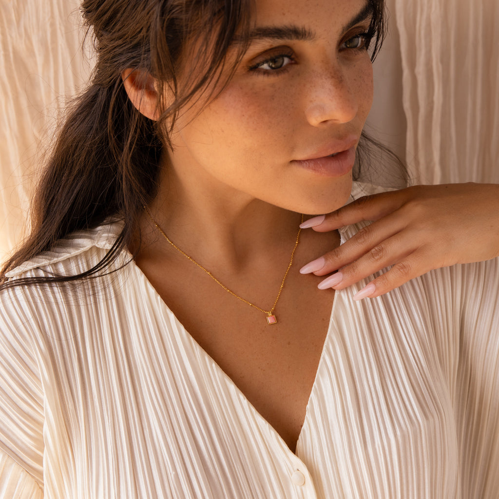 A woman in a cream pleated blouse gently touches her collarbone while wearing the Pink Opal Pendant Necklace, featuring a gold satellite chain with a pink opal pendant.