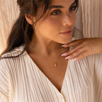 A woman in a cream pleated blouse gently touches her collarbone while wearing the Pink Opal Pendant Necklace, featuring a gold satellite chain with a pink opal pendant.