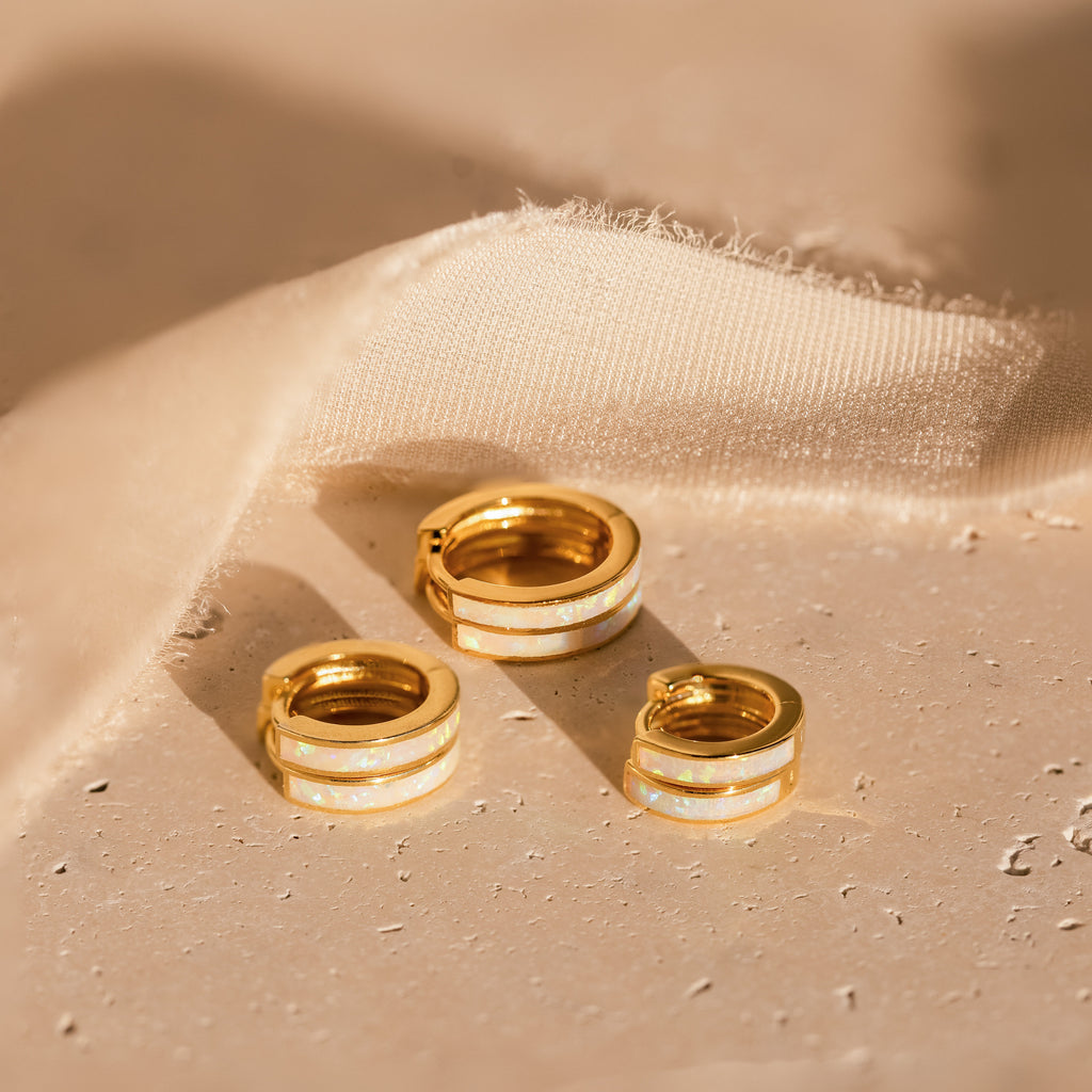 Three White Opal Inlay Huggies are displayed on a light textured surface next to beige fabric.