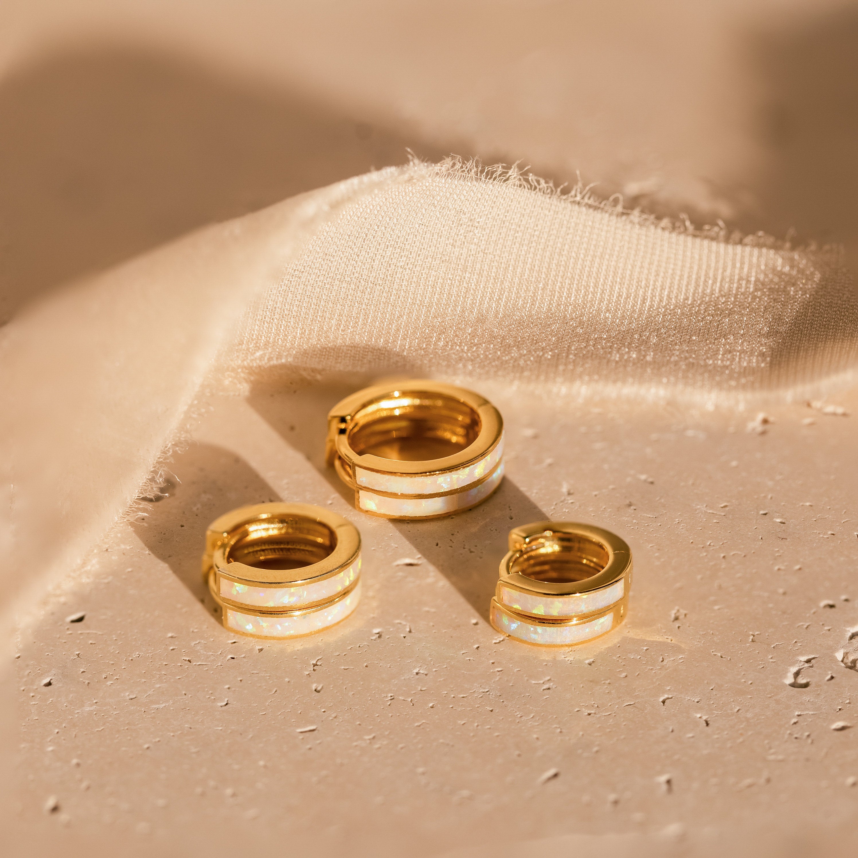 Three White Opal Inlay Huggies are displayed on a light textured surface next to beige fabric.