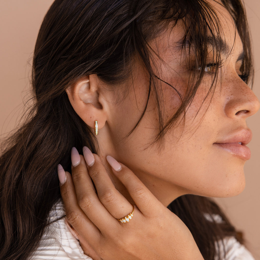 Woman with wavy hair touches her face with manicured nails, wearing gold White Opal Inlay Hoops and a stylish ring.