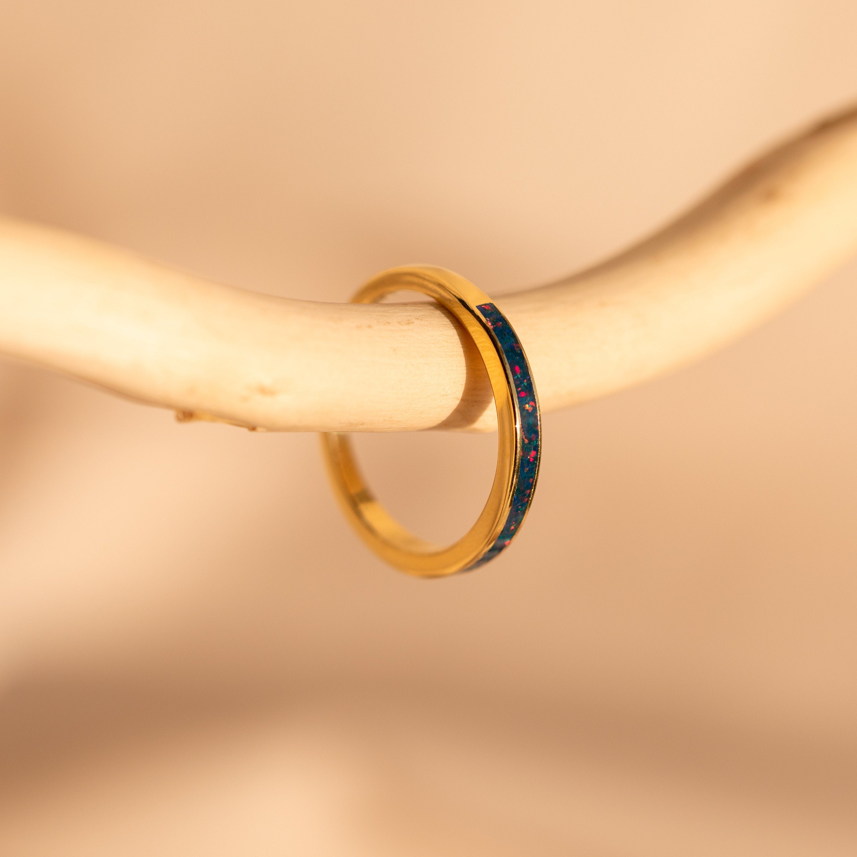 The Indigo Opal Inlay Band, featuring delicate red accents, hangs on a smooth, light branch—ideal for stacking and adding a unique touch to your collection.