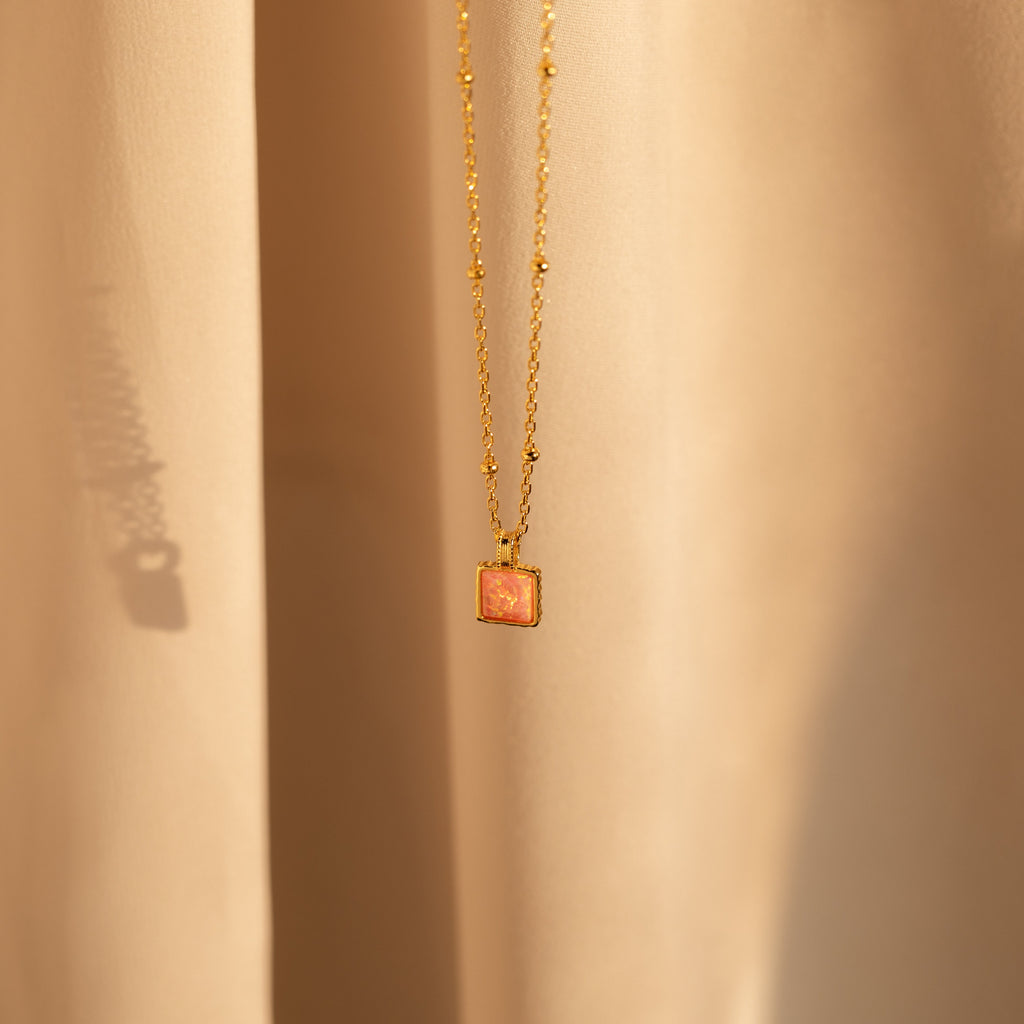 The Pink Opal Pendant Necklace features a gold minimalist design with a square pink opal charm on a satellite chain, displayed against soft beige fabric.