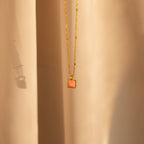 The Pink Opal Pendant Necklace features a gold minimalist design with a square pink opal charm on a satellite chain, displayed against soft beige fabric.