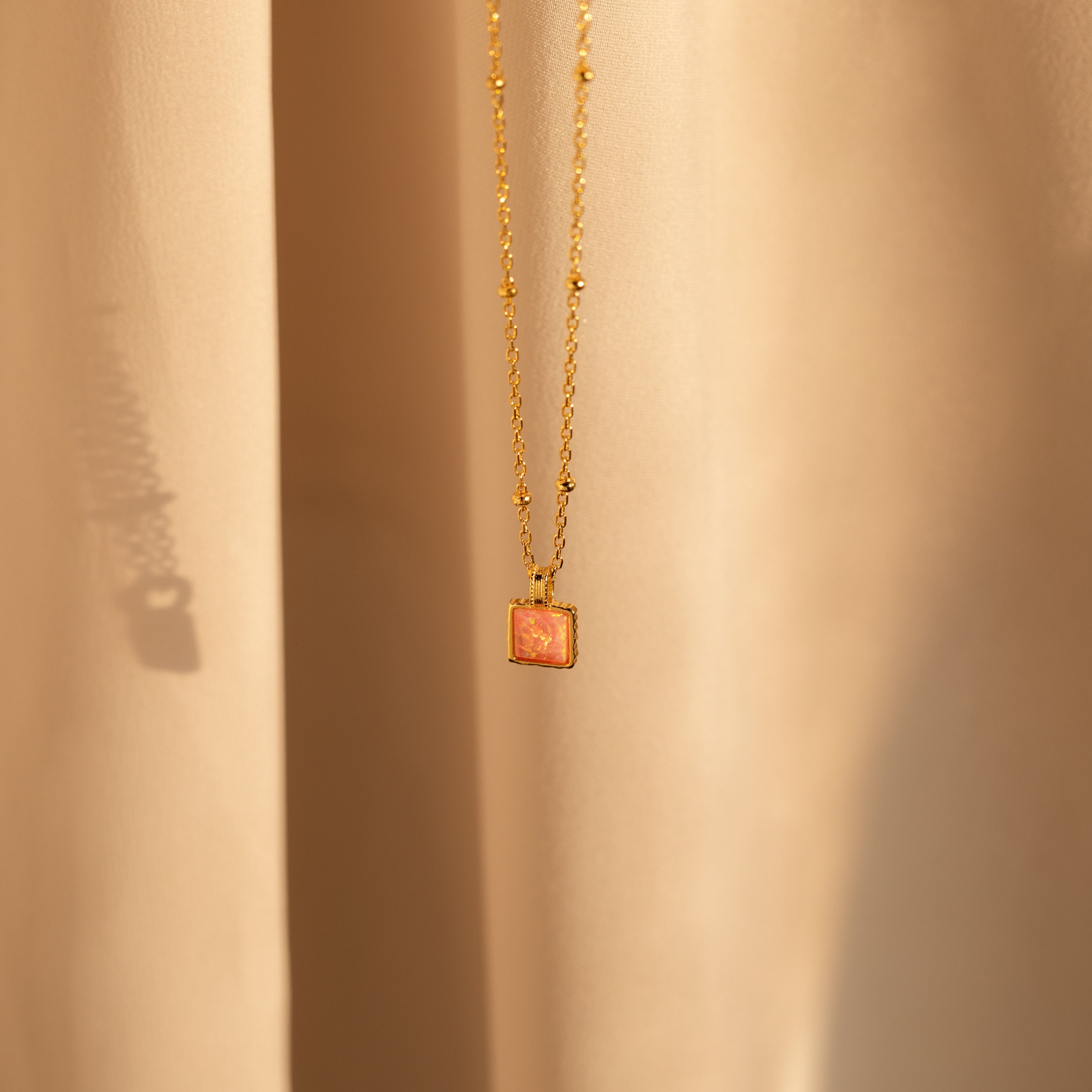 The Pink Opal Pendant Necklace features a gold minimalist design with a square pink opal charm on a satellite chain, displayed against soft beige fabric.