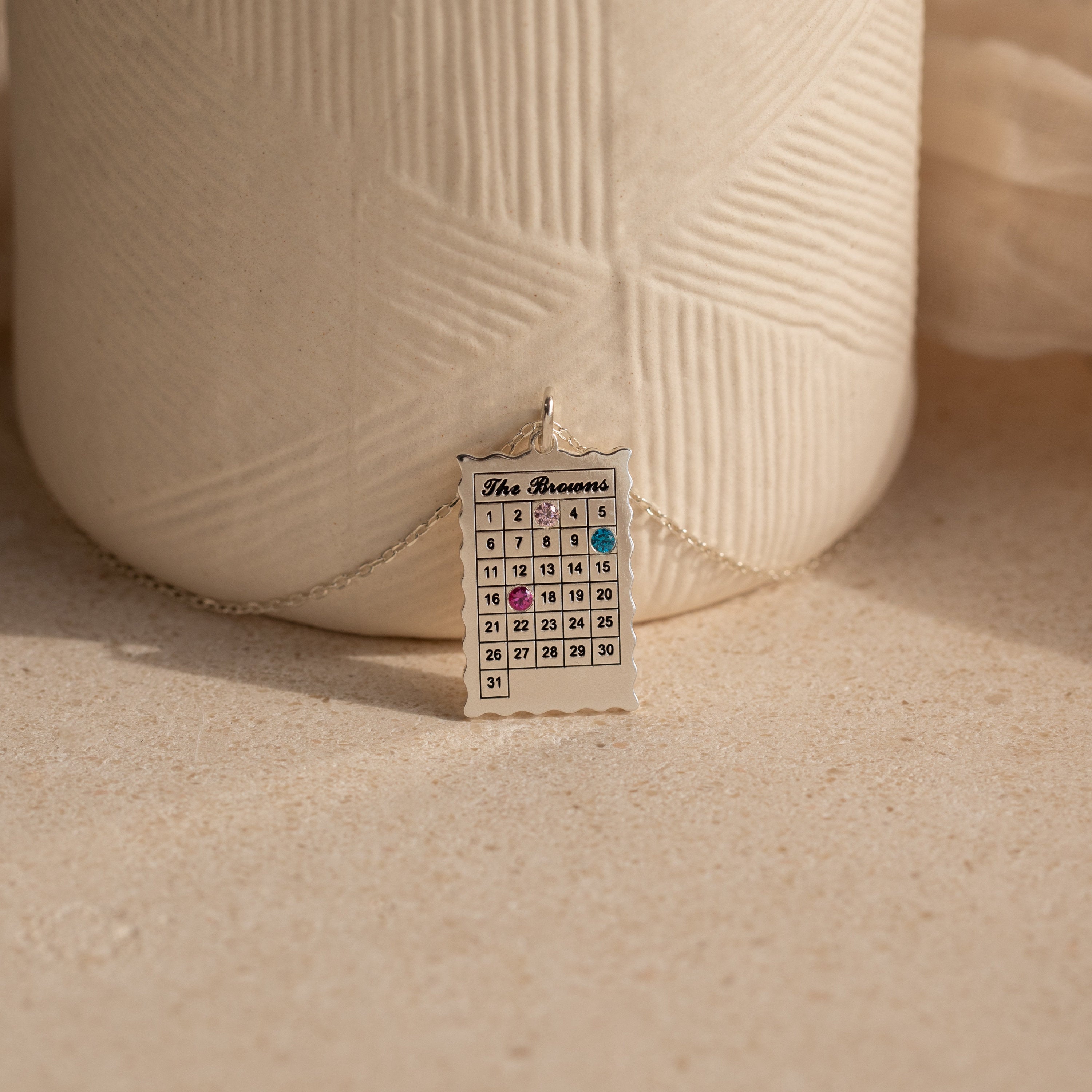 The Calendar Birthstone Necklace features a personalized calendar pendant with highlighted dates, displayed before a textured ceramic vase—a unique and meaningful gift.