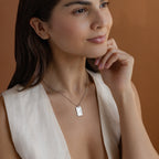 A woman with long brown hair wears a sleeveless cream top and a Floral Name Birthstone Necklace against a warm brown background.