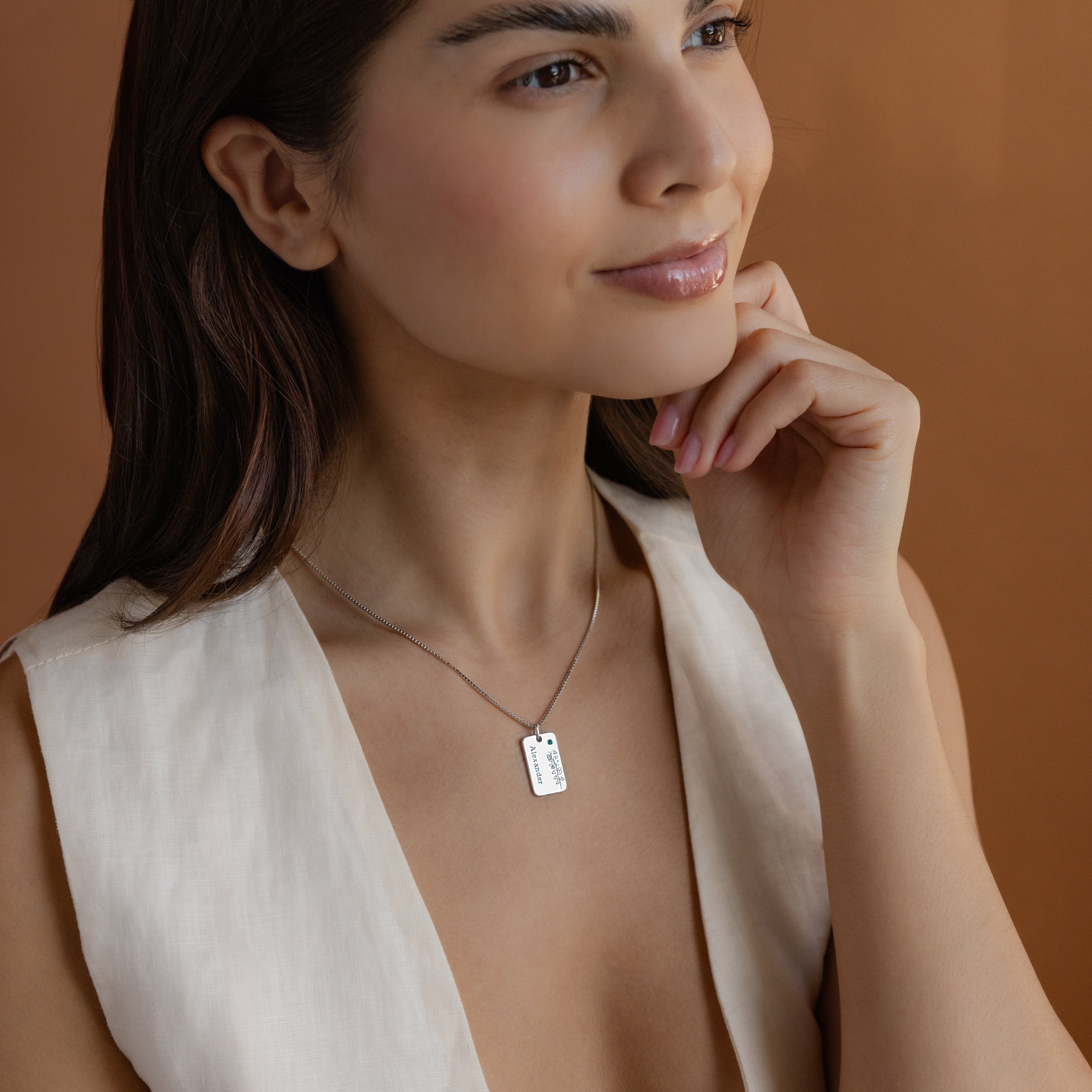 A woman with long brown hair wears a sleeveless cream top and a Floral Name Birthstone Necklace against a warm brown background.