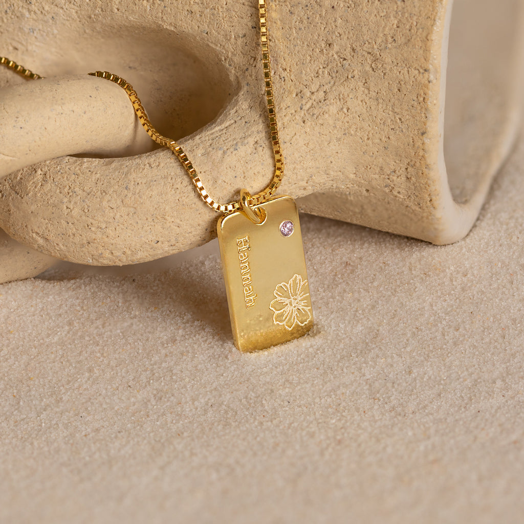 Floral Name Birthstone Necklace featuring a rectangular pendant engraved with "Hanalei," a delicate floral design, and a small gemstone—an elegant birthstone necklace shown on a stone surface.