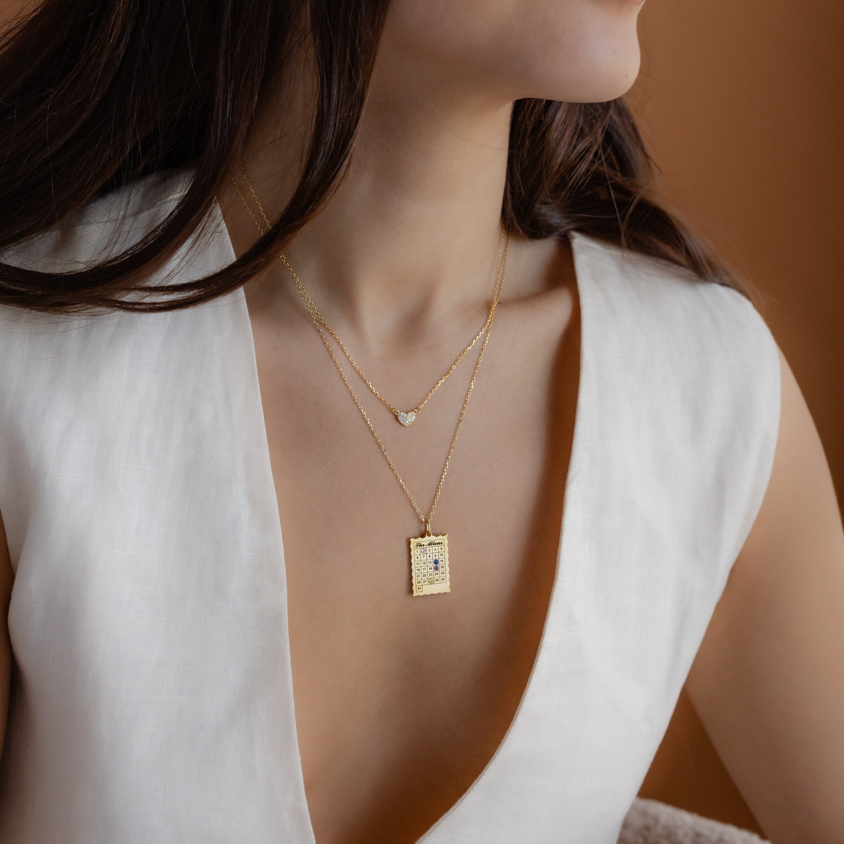 Woman wearing a white V-neck dress and layered gold necklaces, featuring the Calendar Birthstone Necklace, against a brown background.