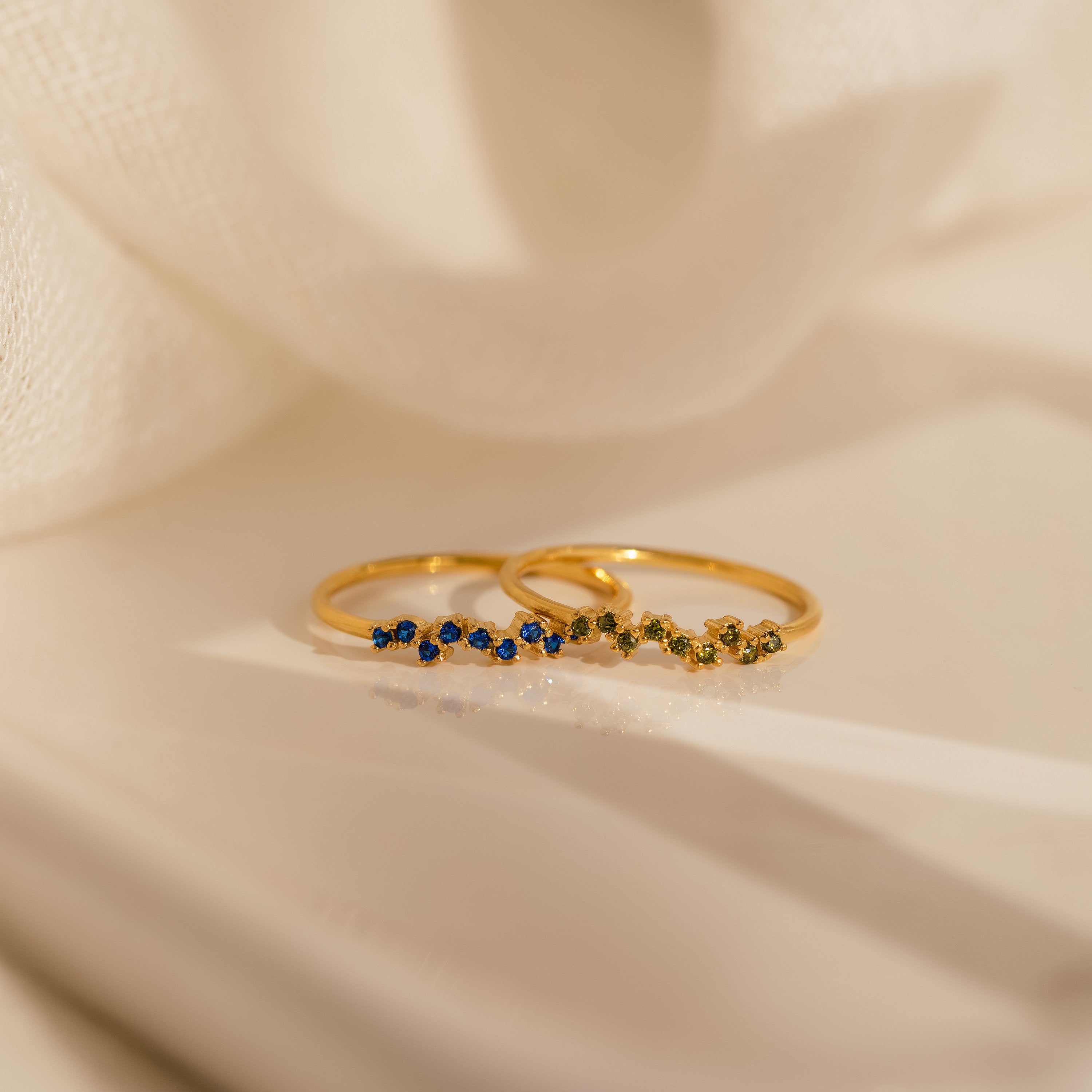 The Tiny Cluster Birthstone Ring features two delicate gold stackable bands with small blue and green gemstones, beautifully displayed on a soft background—ideal as a customizable birthstone ring or worn together for a charming look.