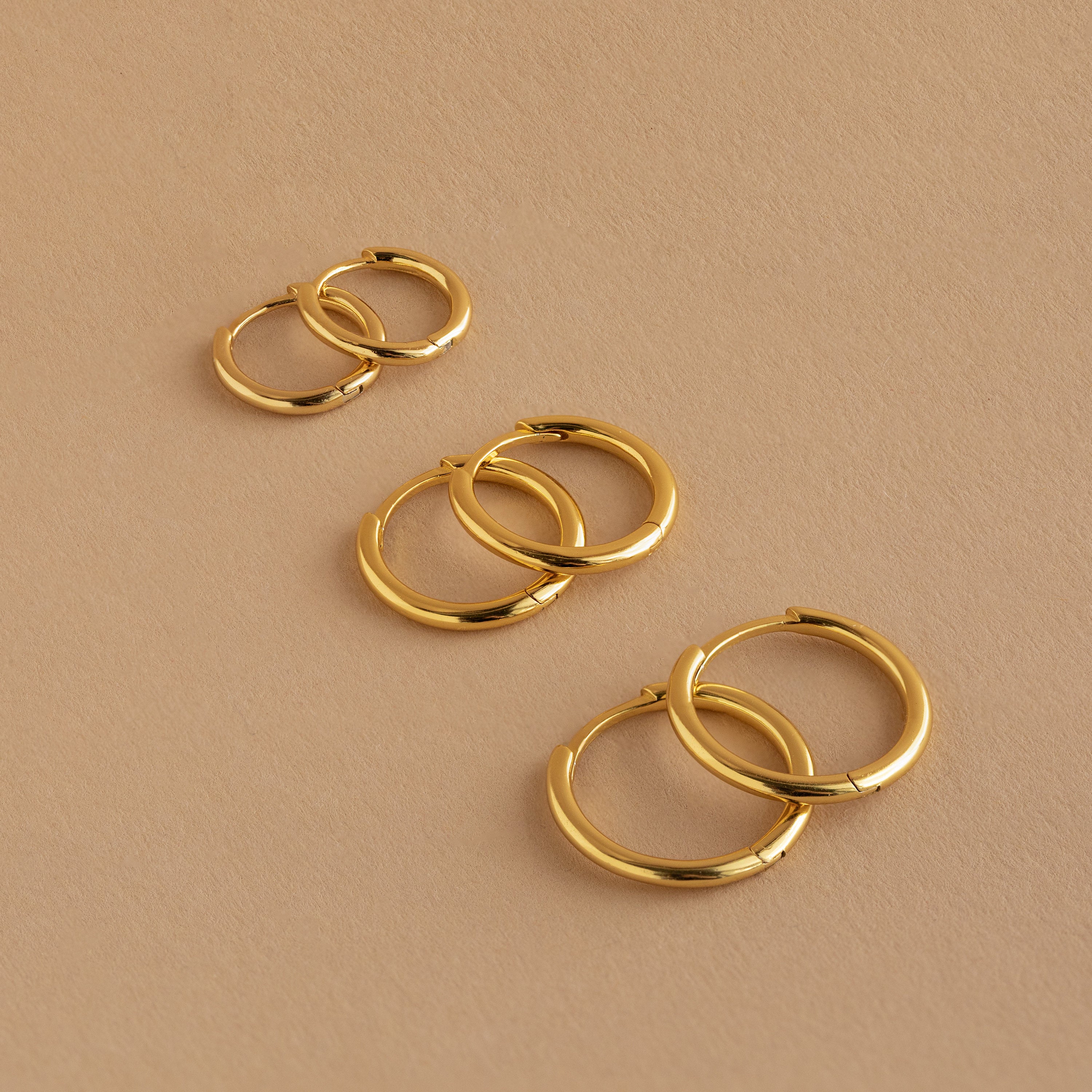 The Mini Bold Hoops Set—three pairs of classic hoops in small, medium, and large—are arranged on a beige surface.