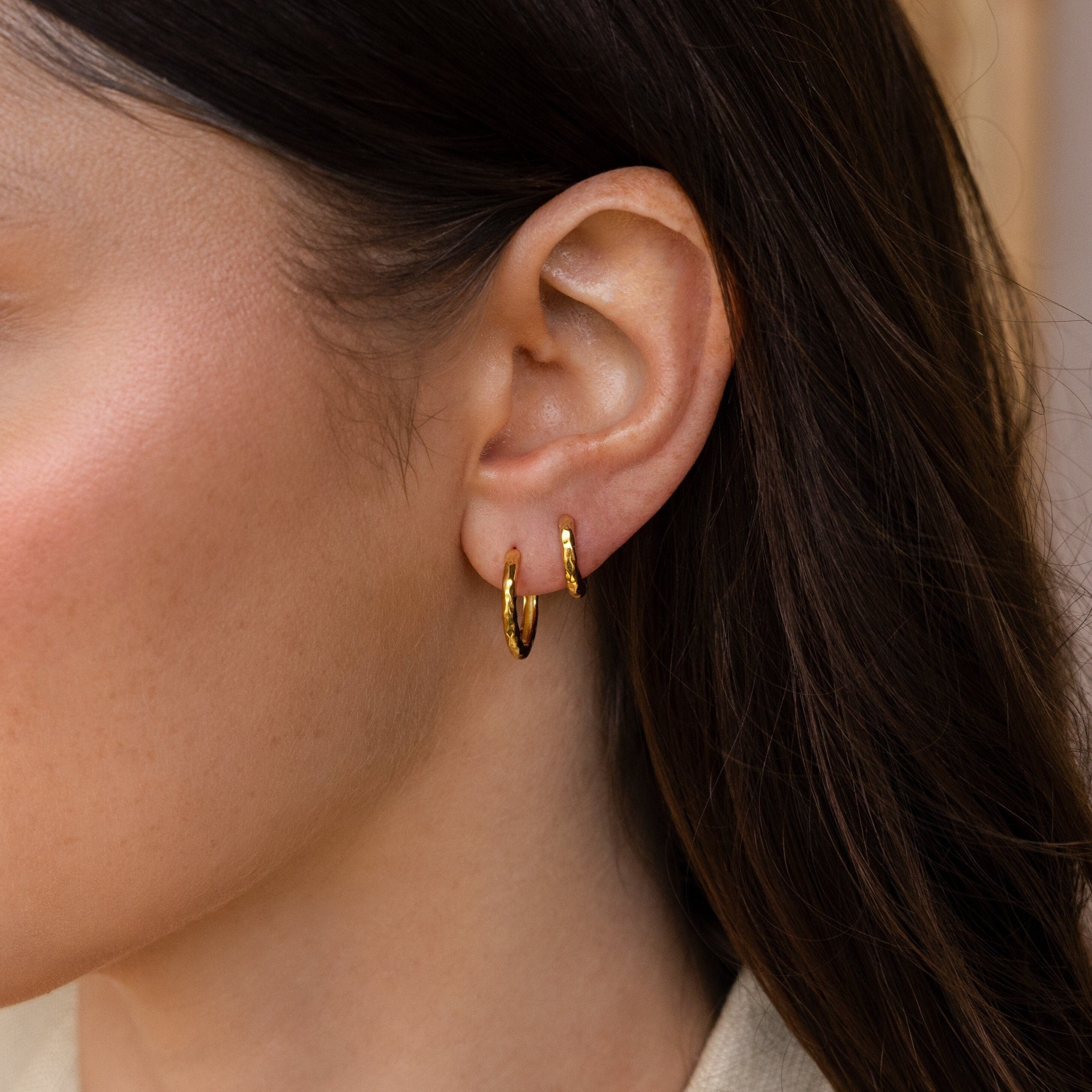 Close-up of a woman's ear with two Mini Hammered Hoops and long brown hair, highlighting the elegance of these gold or silver hoop earrings.
