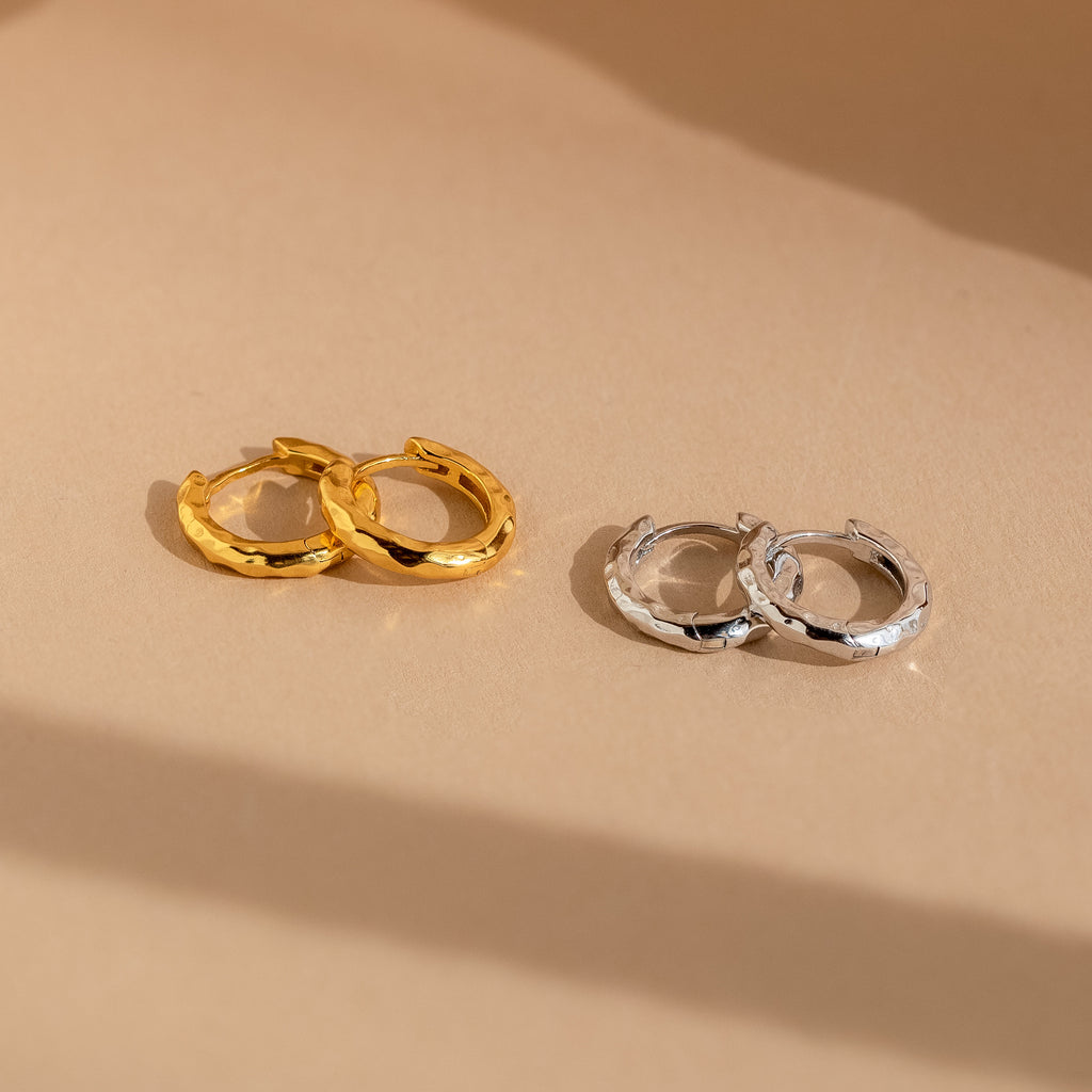The Mini Hammered Hoops Set features gold and silver hammered hoop earrings displayed on a beige surface, offering classic hoops in both finishes perfect for any occasion.
