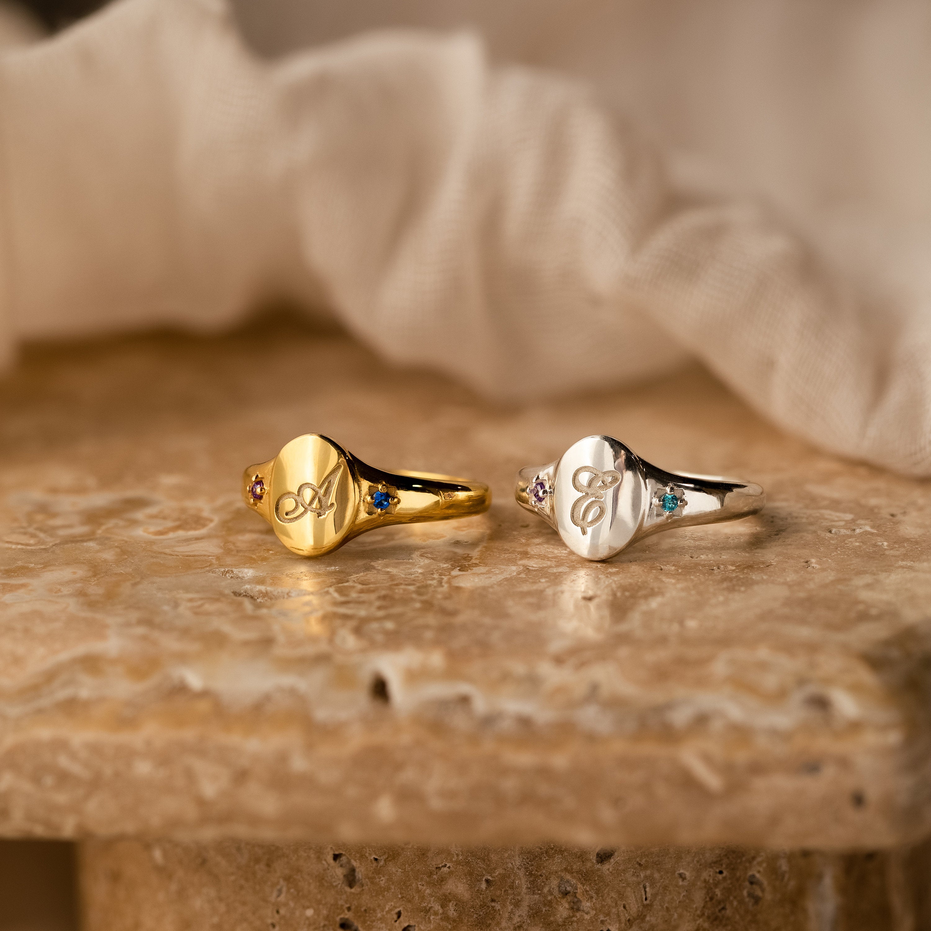 Two engraved Initial Birthstone Signet Rings—one gold, one silver—with colored gemstones, displayed on stone. Personalize these rings for a unique custom gift or stylish accessory for any occasion.