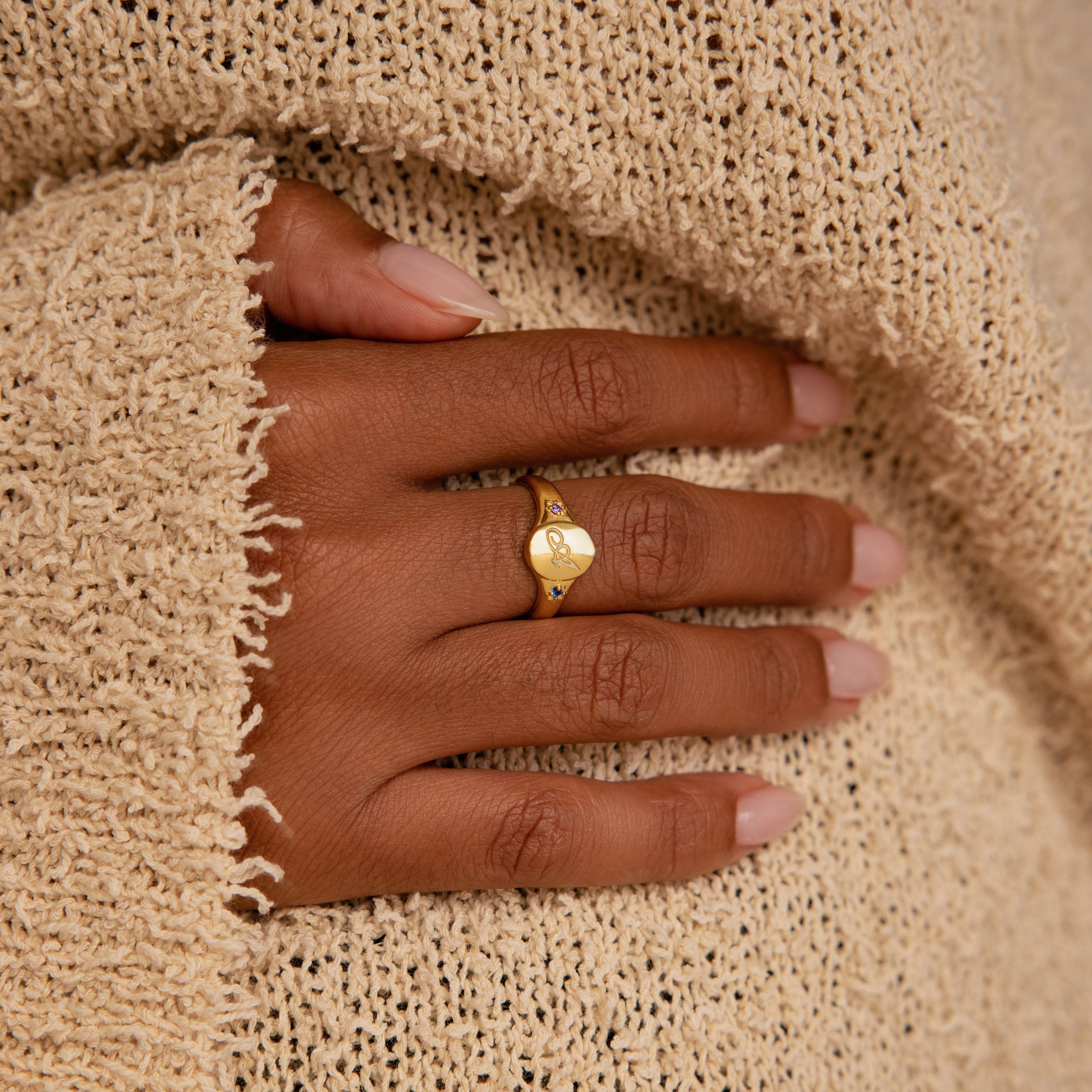 A hand with manicured nails wears the Initial Birthstone Signet Ring, resting on a textured beige sweater.