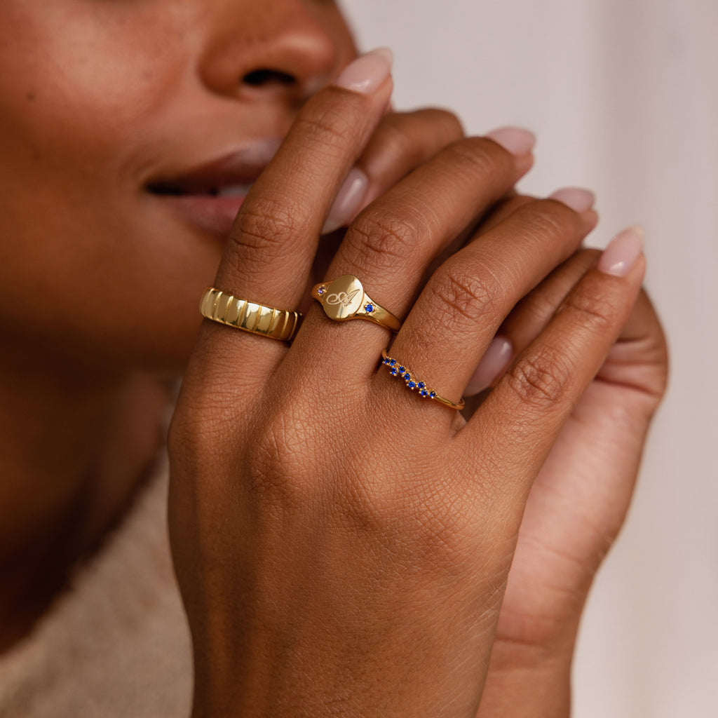 A close-up of a woman's hand showcases three stylish gold rings, featuring the elegant Initial Birthstone Signet Ring, with her nails neatly manicured.