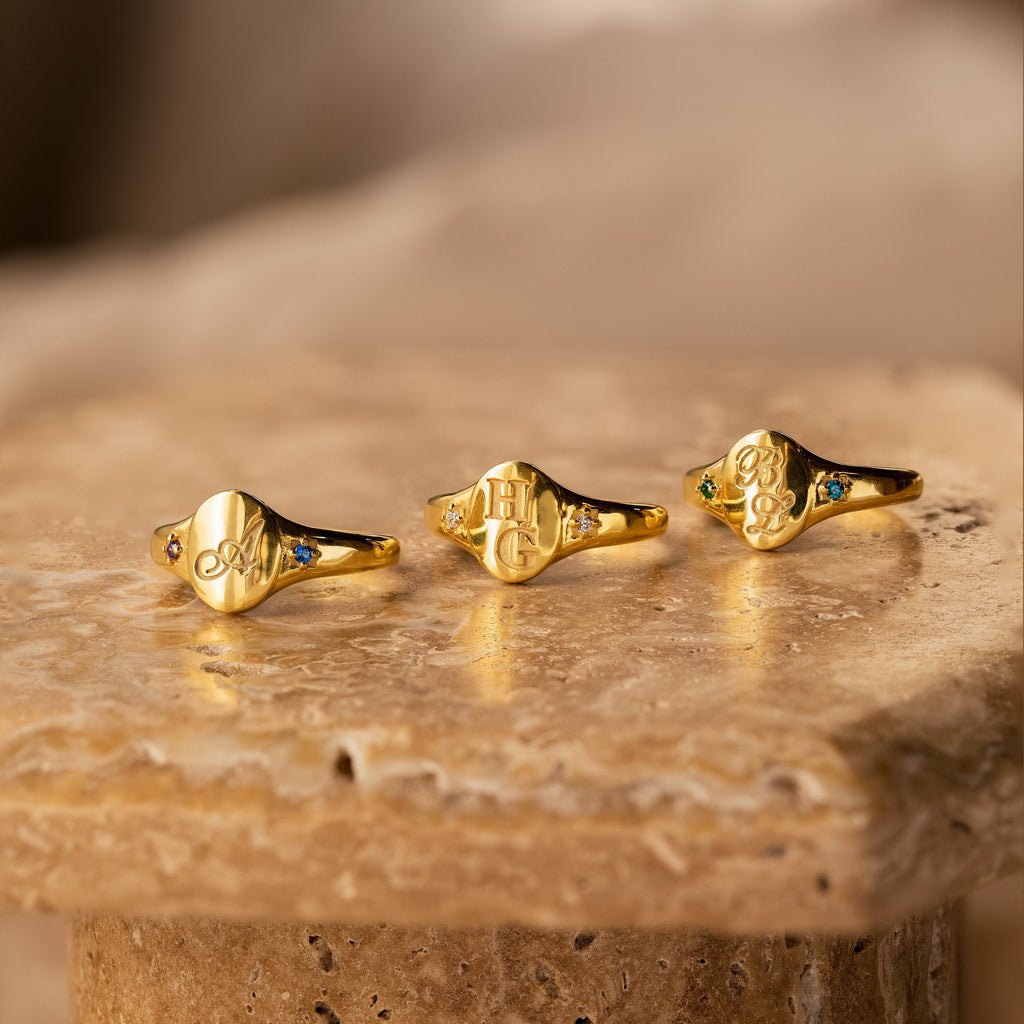 Three gold Initial Birthstone Signet Rings with engraved initials and vibrant gemstones are displayed on a textured stone surface—the perfect personalized piece and custom gift.