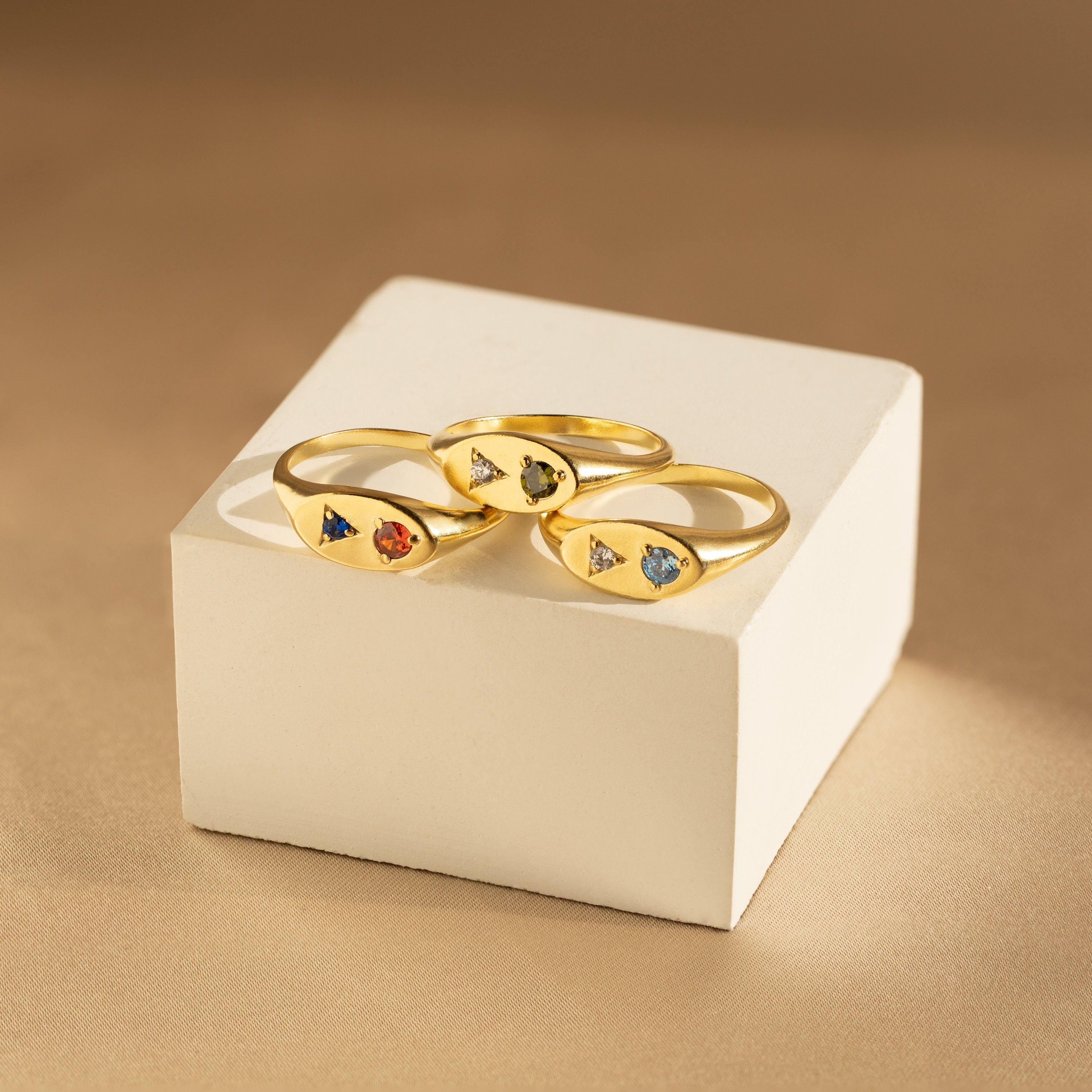 The Mixed Birthstone Signet Rings, featuring vibrant gemstones, are displayed on a white box against a beige background, showcasing the charm of personalized jewelry.