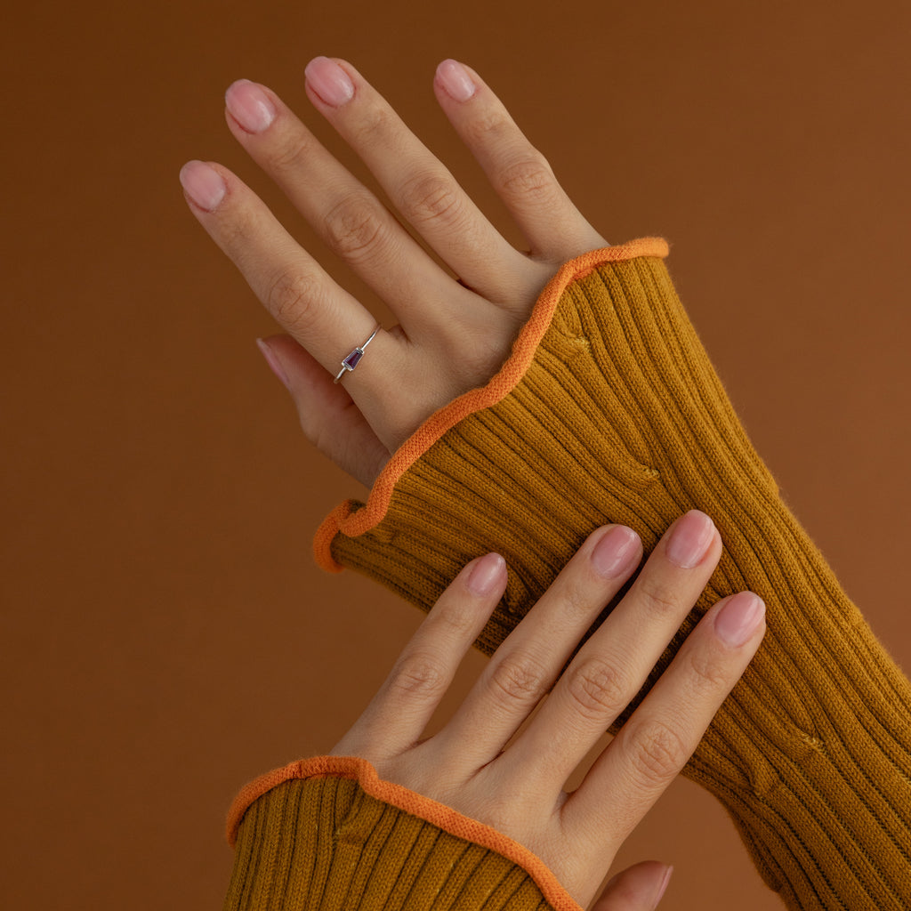 Hands with light pink nails wear the Irregular Baguette Birthstone Ring, paired with mustard ribbed sleeves, set against a brown background.