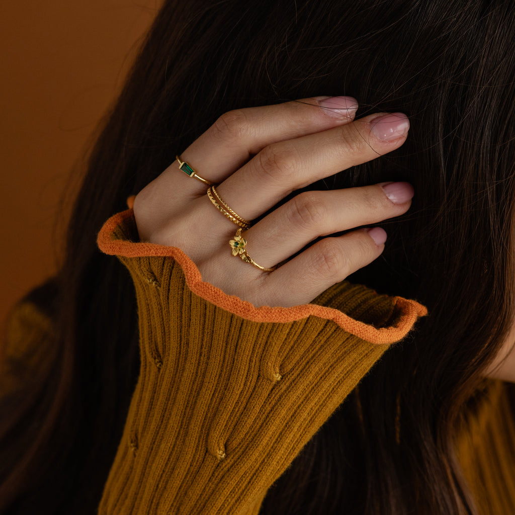 A hand with pink nails wearing the Irregular Baguette Birthstone Ring touches dark hair, paired with a mustard sweater featuring orange trim.