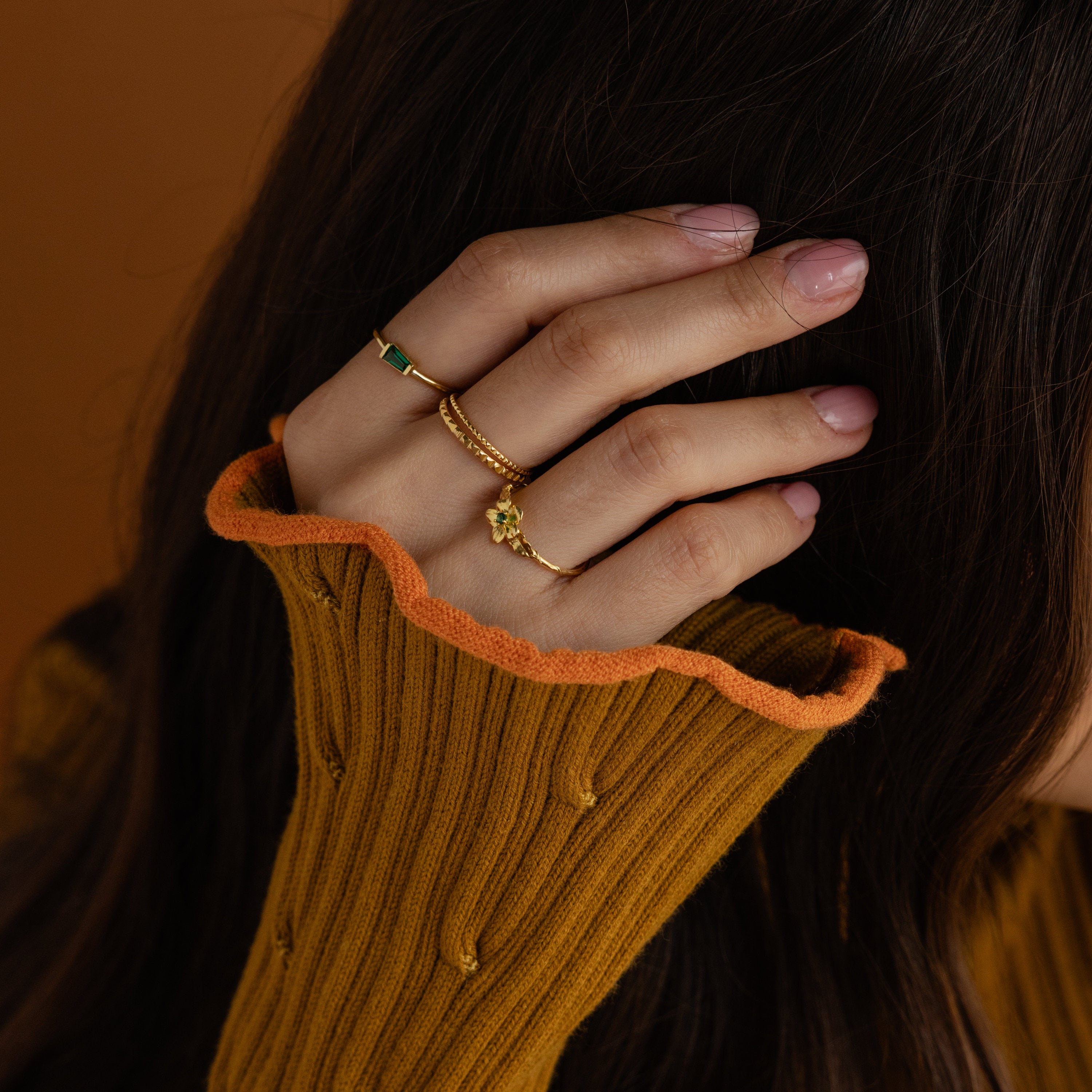 A hand with pink nails wearing the Irregular Baguette Birthstone Ring touches dark hair, paired with a mustard sweater featuring orange trim.