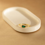 The Irregular Baguette Birthstone Ring, featuring a green rectangular gemstone set in gold, rests on a white oval dish against a beige background.