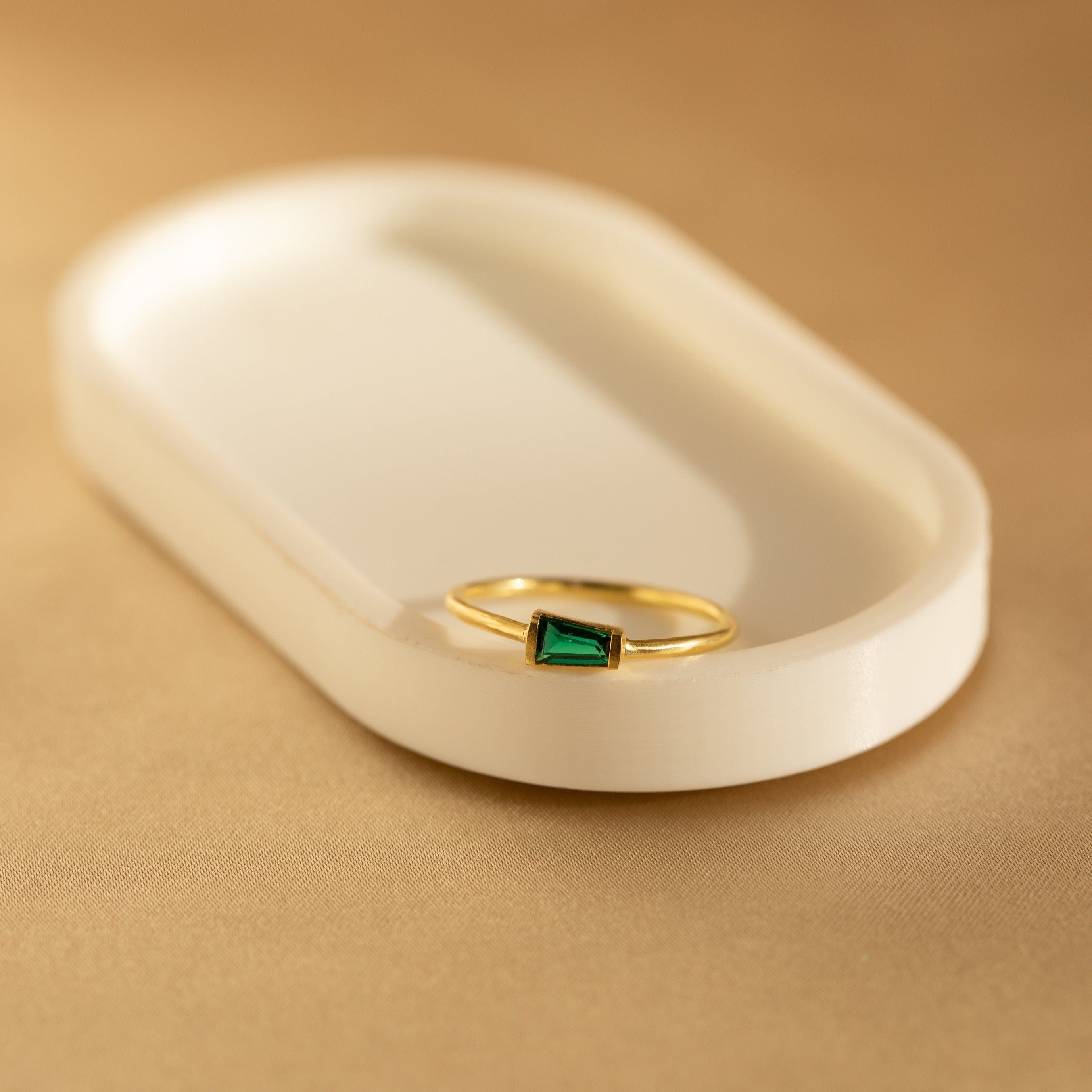 The Irregular Baguette Birthstone Ring, featuring a green rectangular gemstone set in gold, rests on a white oval dish against a beige background.