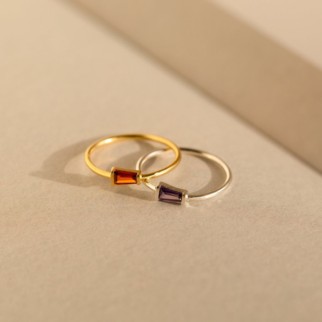 Two Irregular Baguette Birthstone Rings—one gold with a red stone, one silver with a purple stone—rest on a neutral surface. These modern rings are chic accents for stacking or wearing as birthstone jewelry.
