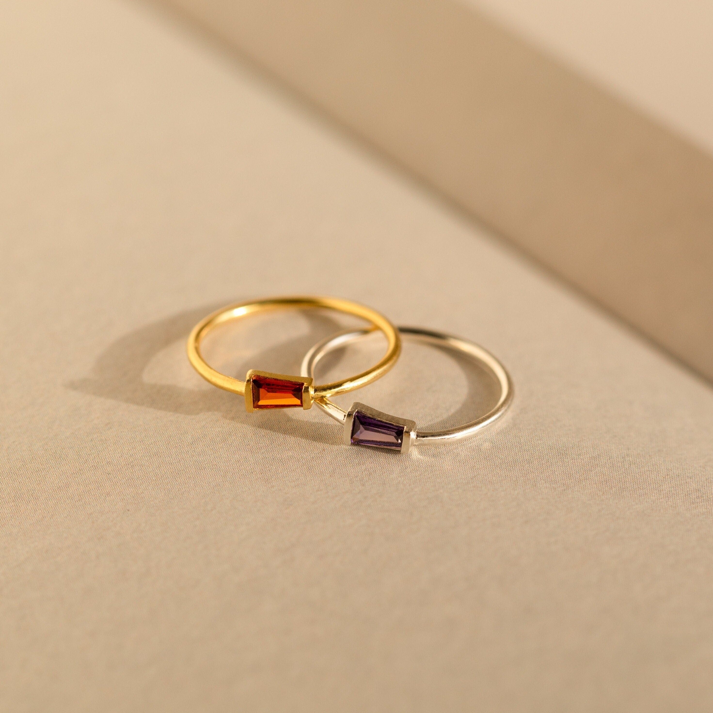 Two Irregular Baguette Birthstone Rings—one gold with a red stone, one silver with a purple stone—rest on a neutral surface. These modern rings are chic accents for stacking or wearing as birthstone jewelry.
