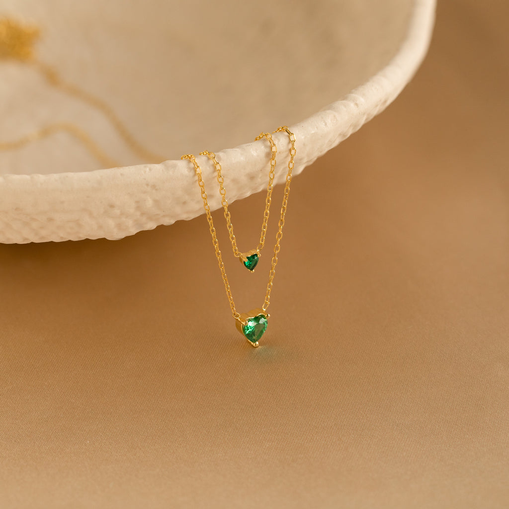 The Heart Birthstone Necklace features two green heart-shaped pendants, elegantly displayed on a textured ceramic bowl atop a beige surface—an ideal personalized jewelry gift for birthdays.