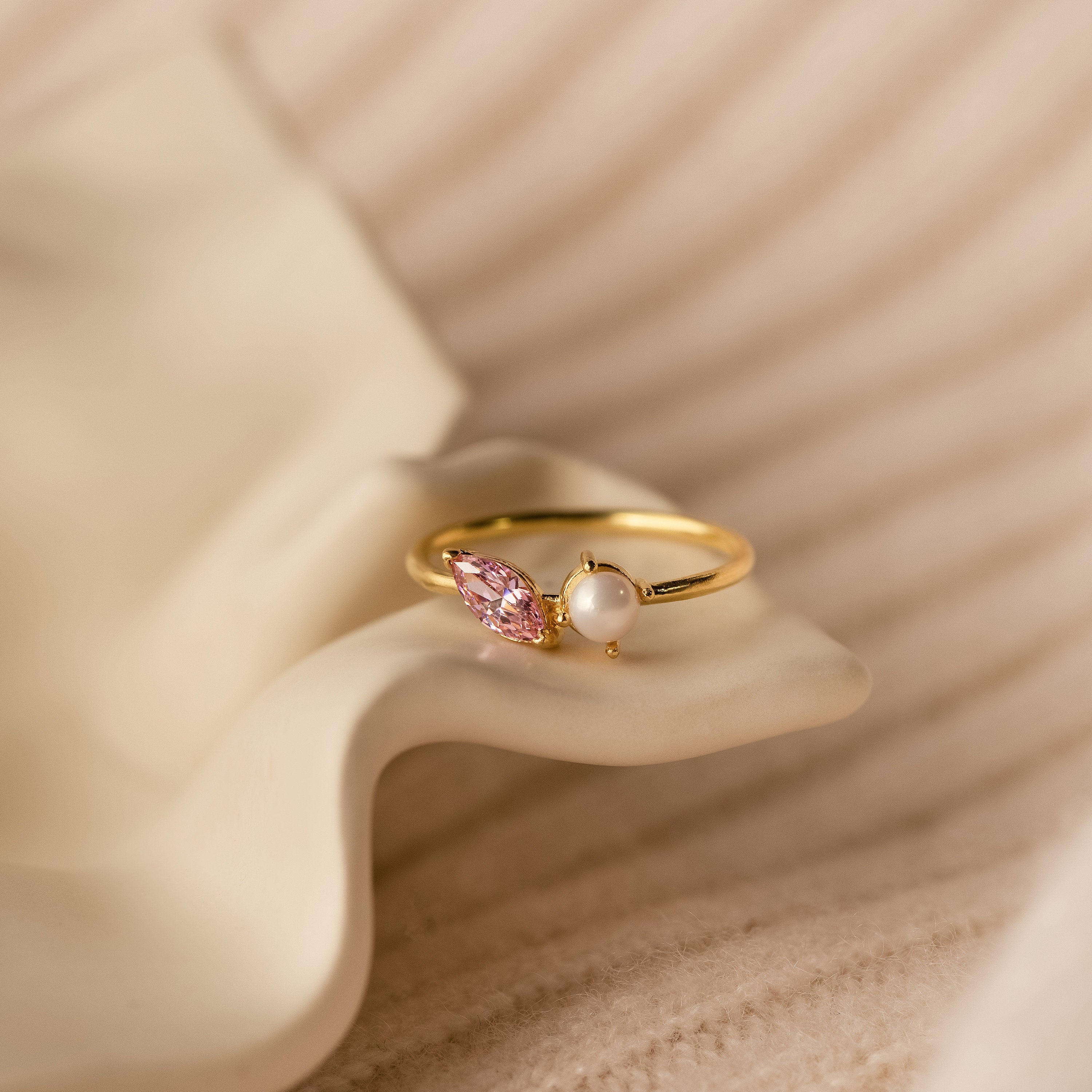 The Marquise Birthstone & Pearl Ring features a pink marquise gem and a pearl, shown stacked on a soft, wavy cream-colored surface.