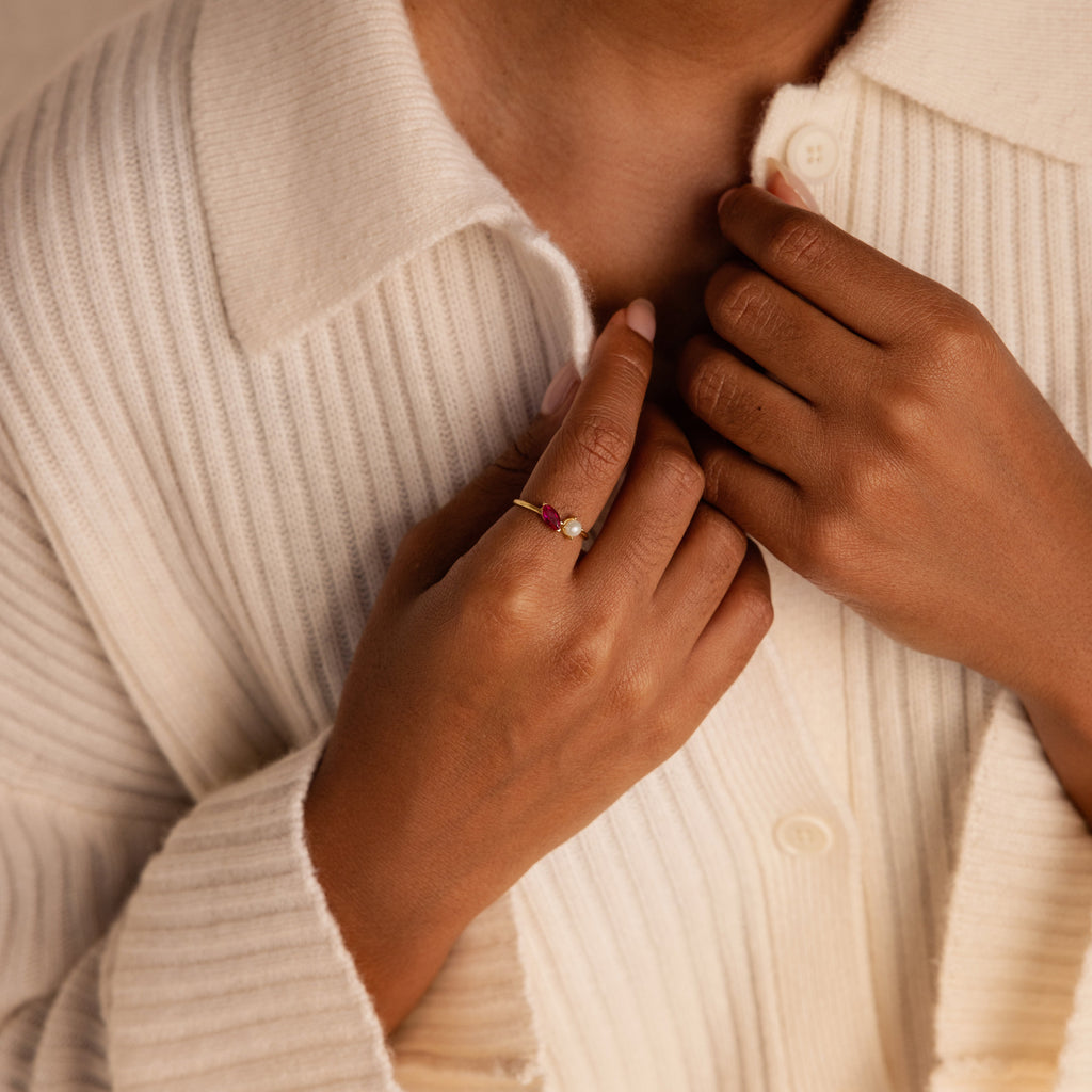 A person wearing a cream ribbed shirt displays the Marquise Birthstone & Pearl Ring, a gold stackable ring with pink stones, on their hand.