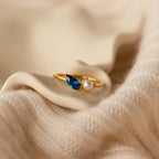 The Marquise Birthstone & Pearl Ring, featuring a blue gem and a pearl, rests on soft, cream-colored fabric.