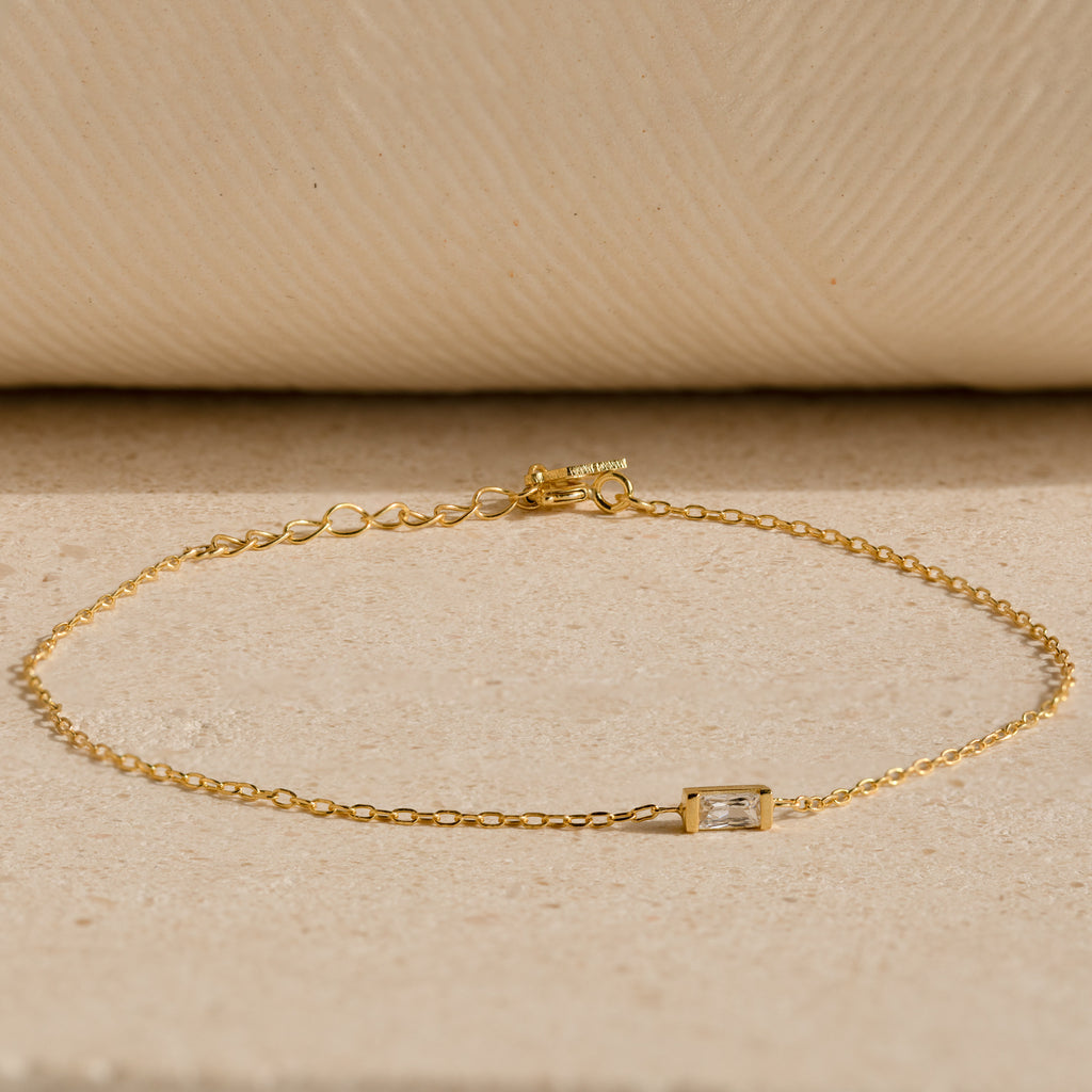 The Baguette Birthstone Bracelet features a delicate gold chain and a single rectangular clear gemstone, shown on a neutral textured surface.