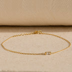 The Baguette Birthstone Bracelet features a delicate gold chain and a single rectangular clear gemstone, shown on a neutral textured surface.