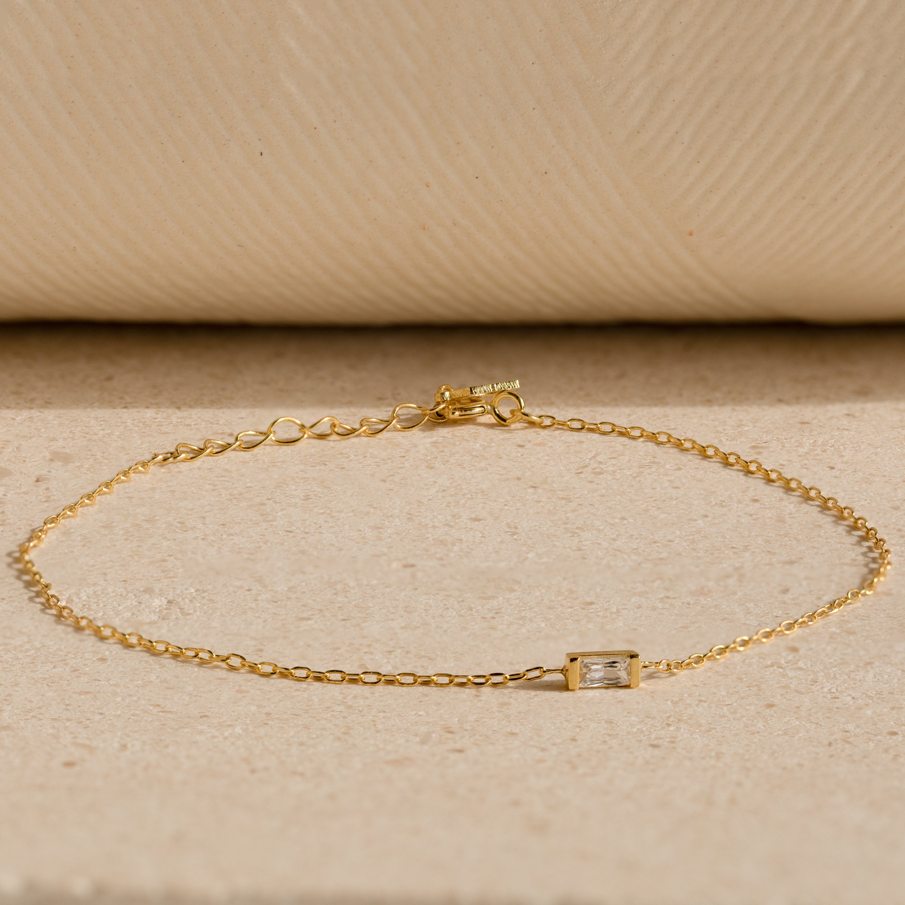 The Baguette Birthstone Bracelet features a delicate gold chain and a single rectangular clear gemstone, shown on a neutral textured surface.