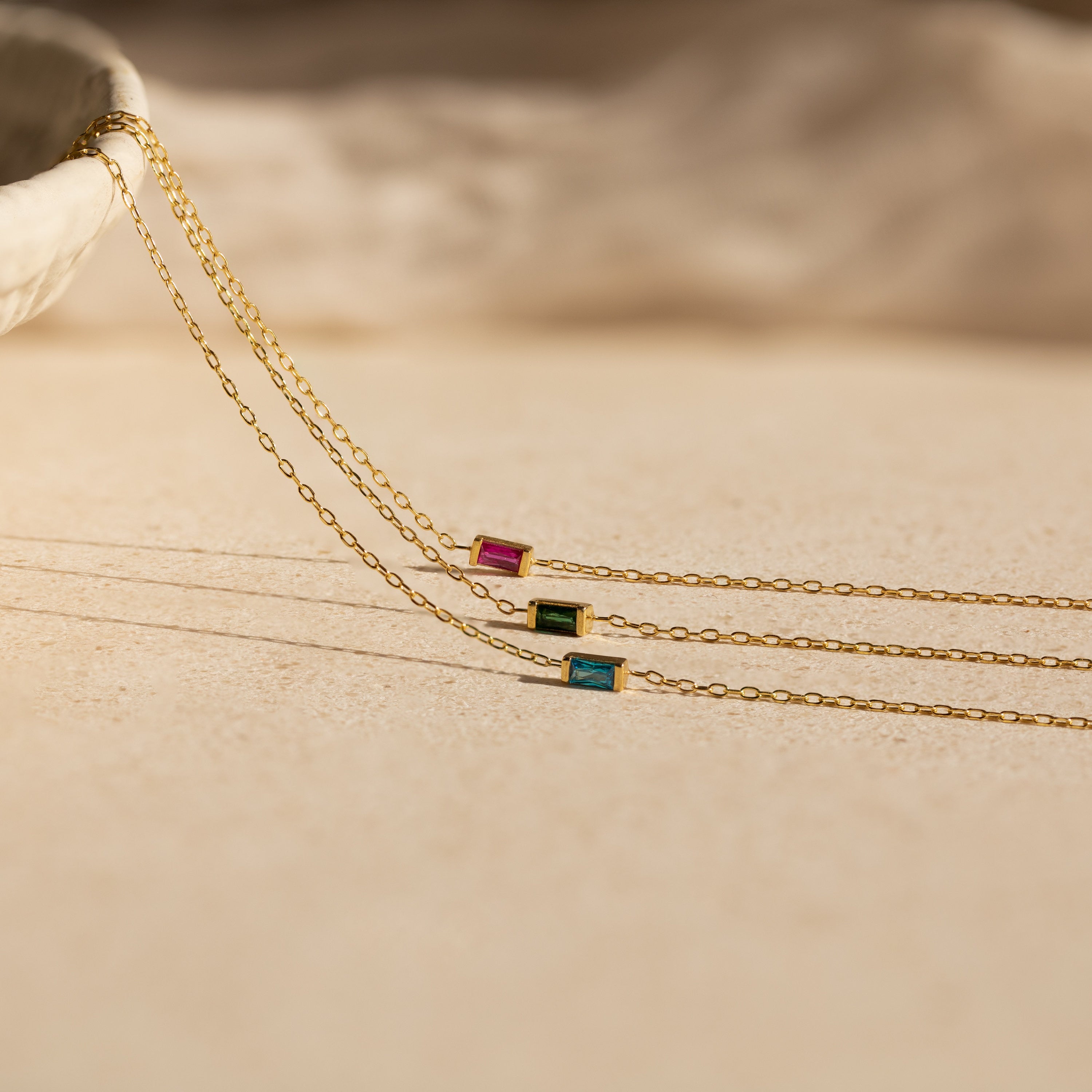 Three delicate gold chain necklaces with red, green, and blue rectangular gemstone pendants rest on a beige surface—ideal for pairing with a personalized bracelet or stacking alongside a Baguette Birthstone Bracelet.