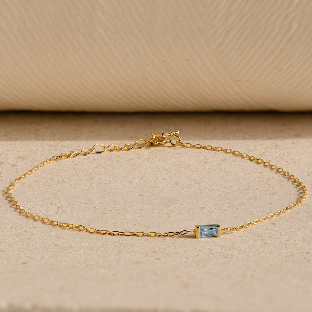The Baguette Birthstone Bracelet features a delicate gold chain and a small rectangular blue gemstone, shown on a beige textured surface.