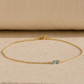 The Baguette Birthstone Bracelet features a delicate gold chain and a small rectangular blue gemstone, shown on a beige textured surface.