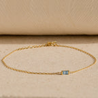 The Baguette Birthstone Bracelet features a delicate gold chain and a small rectangular blue gemstone, shown on a beige textured surface.